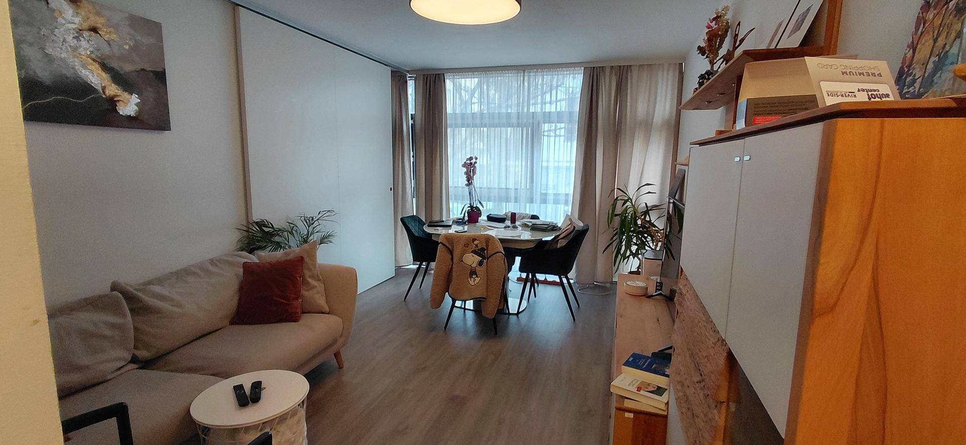 Heller Wohn- und Essbereich mit Holzboden, beigem Sofa und Esstisch am großen Fenster.