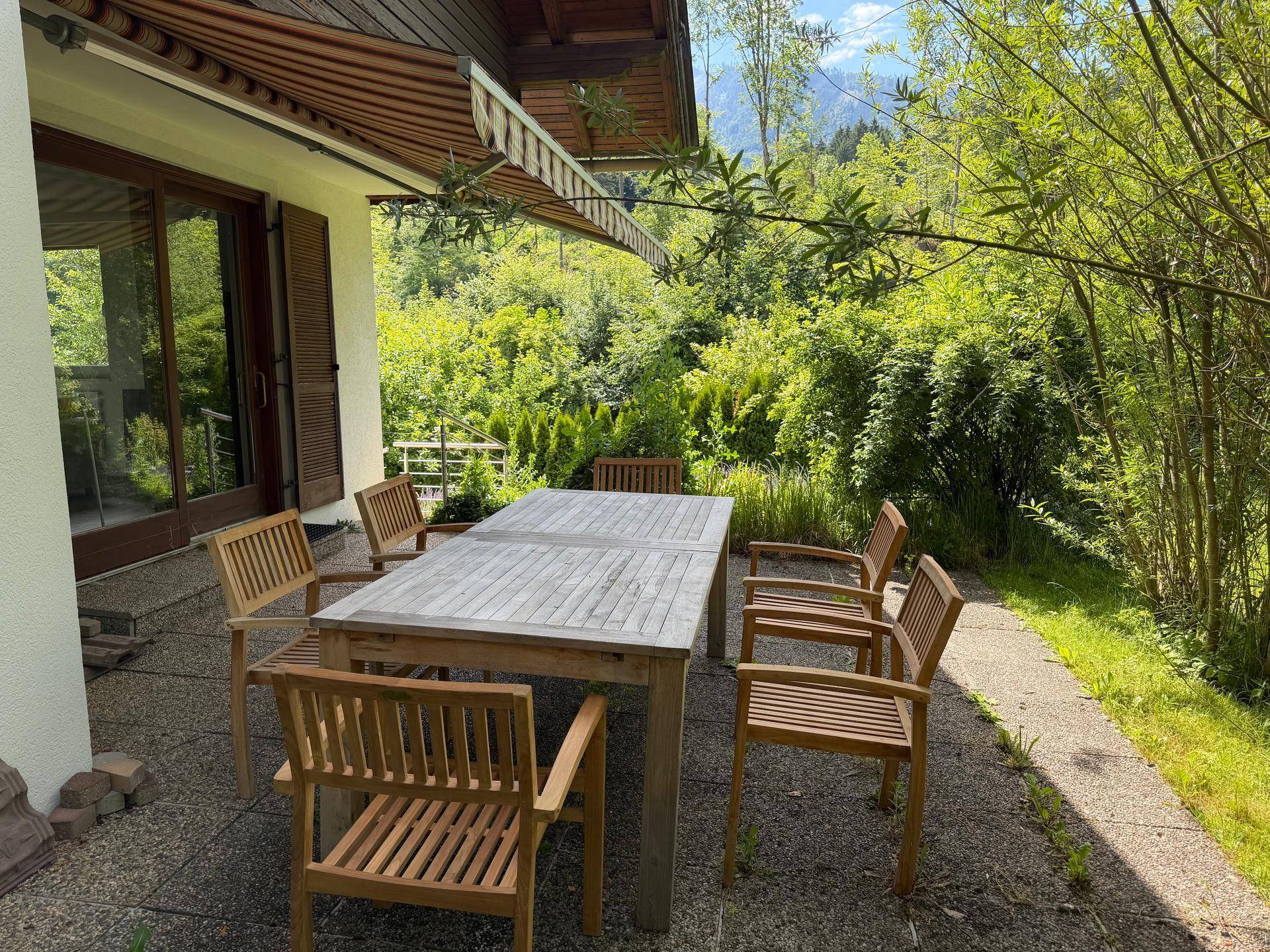 Sonniger Terrassenbereich mit Holztisch und Stühlen, umgeben von üppigem Grün.