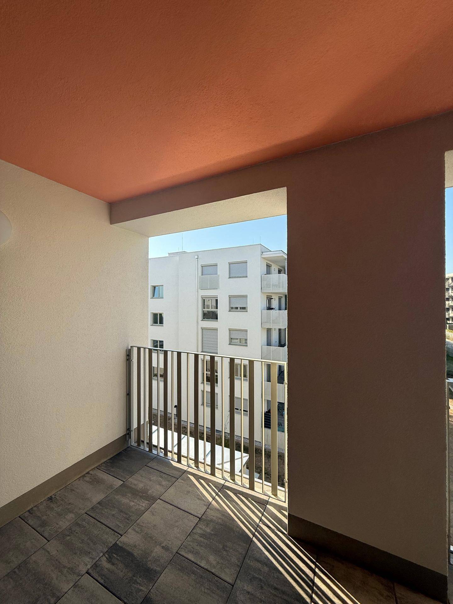 Heller Balkon mit Fliesenboden, Metallgeländer und Blick auf ein modernes Gebäude.
