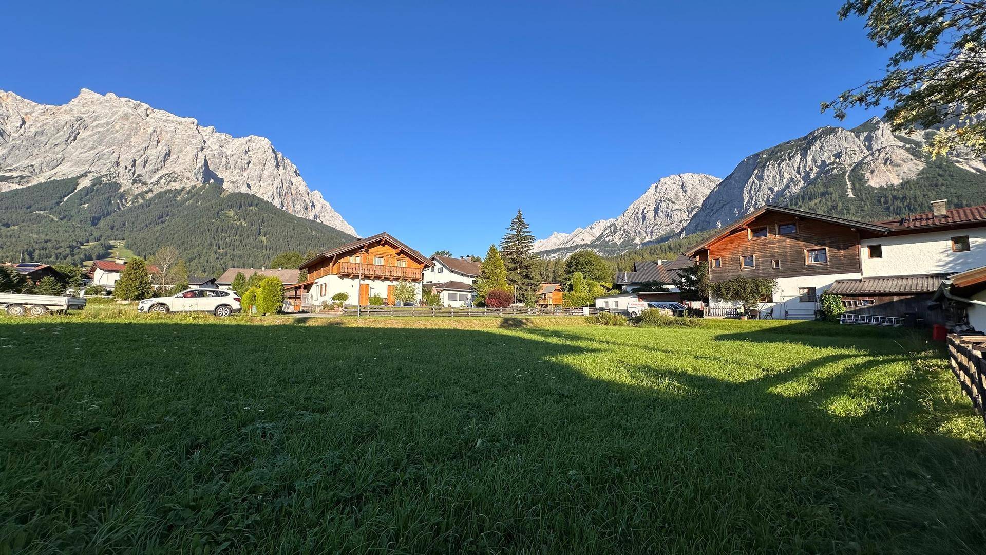 Weitläufige grüne Wiese mit mehreren Häusern und einer beeindruckenden Bergkulisse im Sonnenschein.