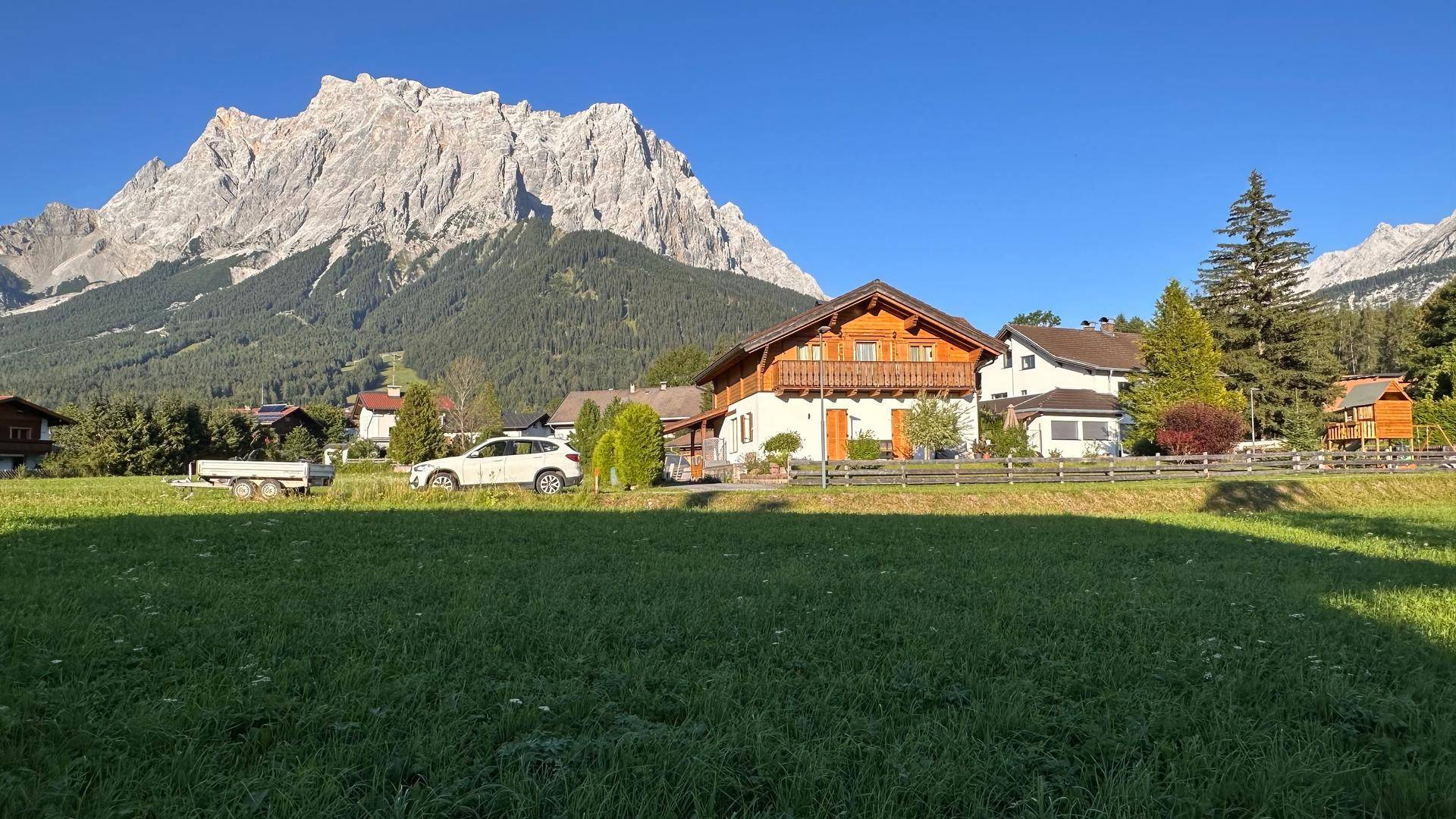 Traditionelles Haus mit Holzbalkonen und einem Anhänger, umgeben von grünen Wiesen und Bergen.