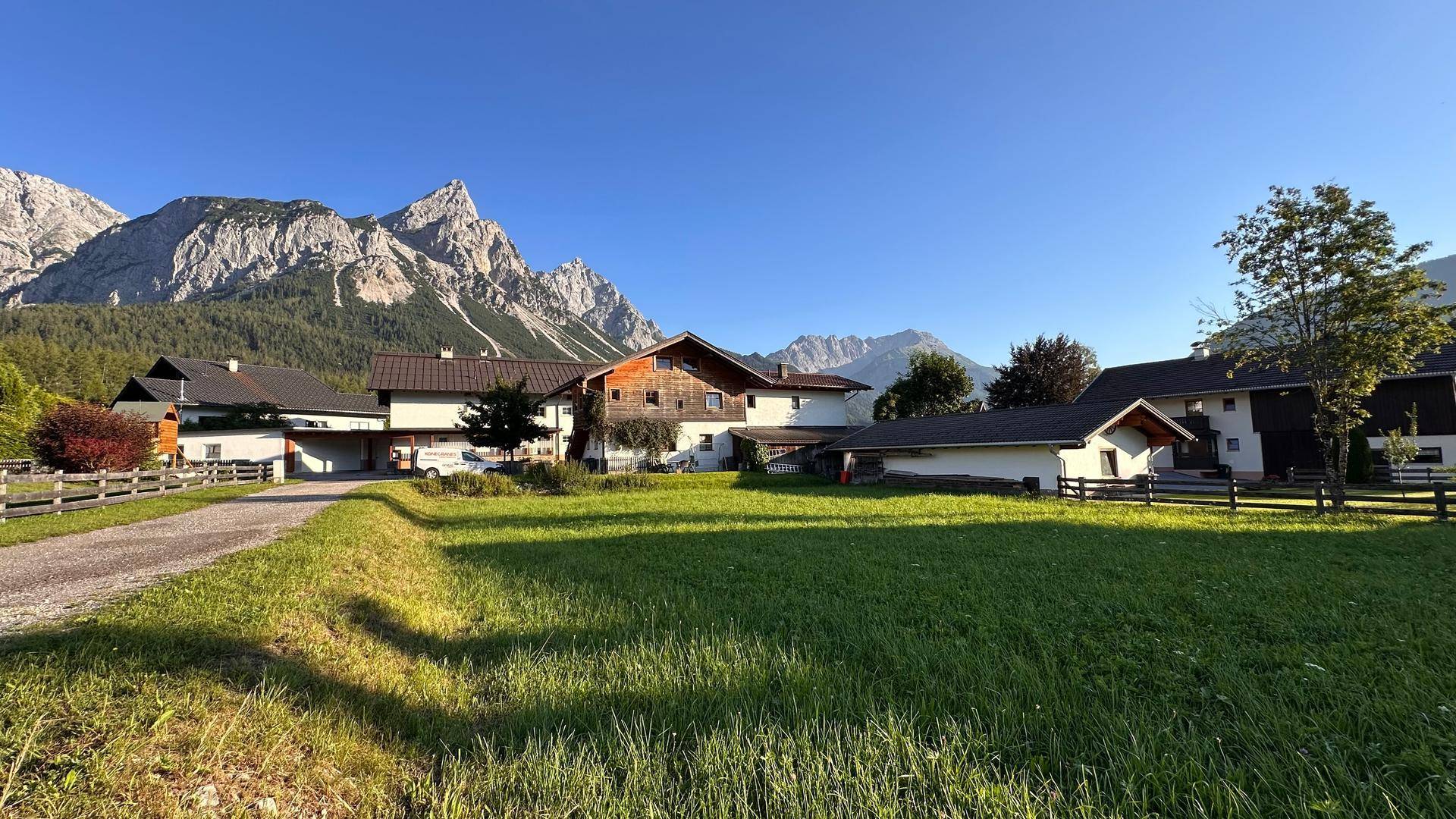 Sonnige Landschaft mit einem Schotterweg, Häusern und einer malerischen Bergkulisse im Hintergrund.