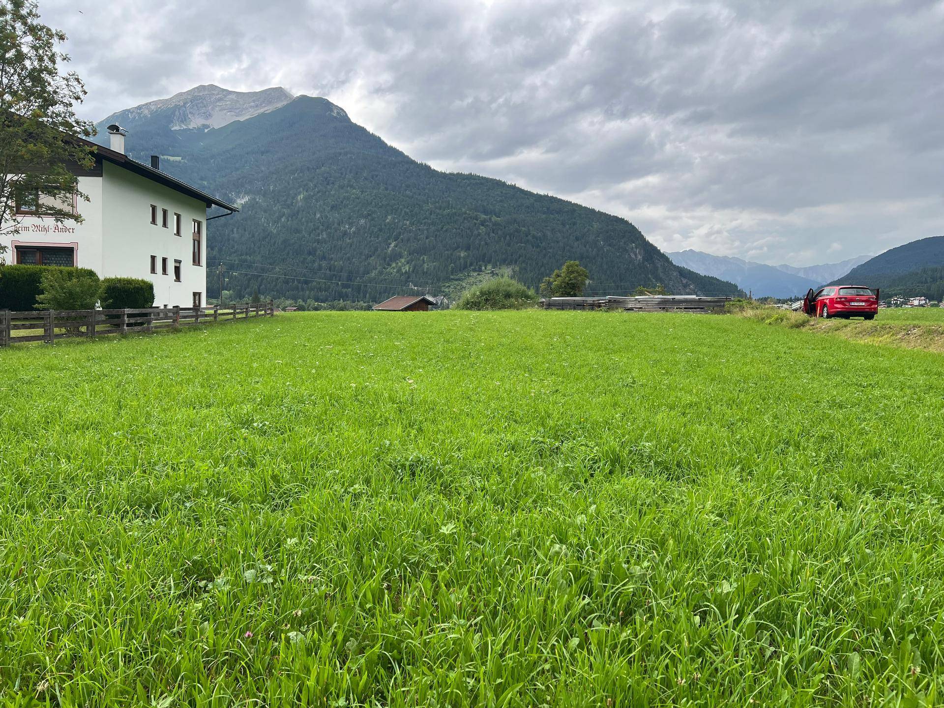 Weitläufige grüne Wiese mit einem Gebäude am Rand und einer Bergkette im Hintergrund.