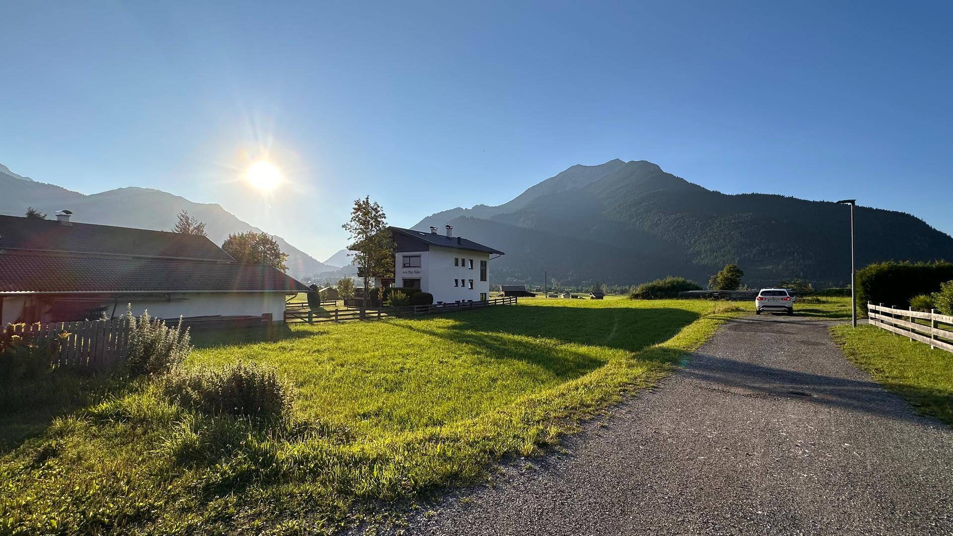 Sonnige Wiese mit einem Schotterweg und einem Wohnhaus, umgeben von einer weiten Berglandschaft.
