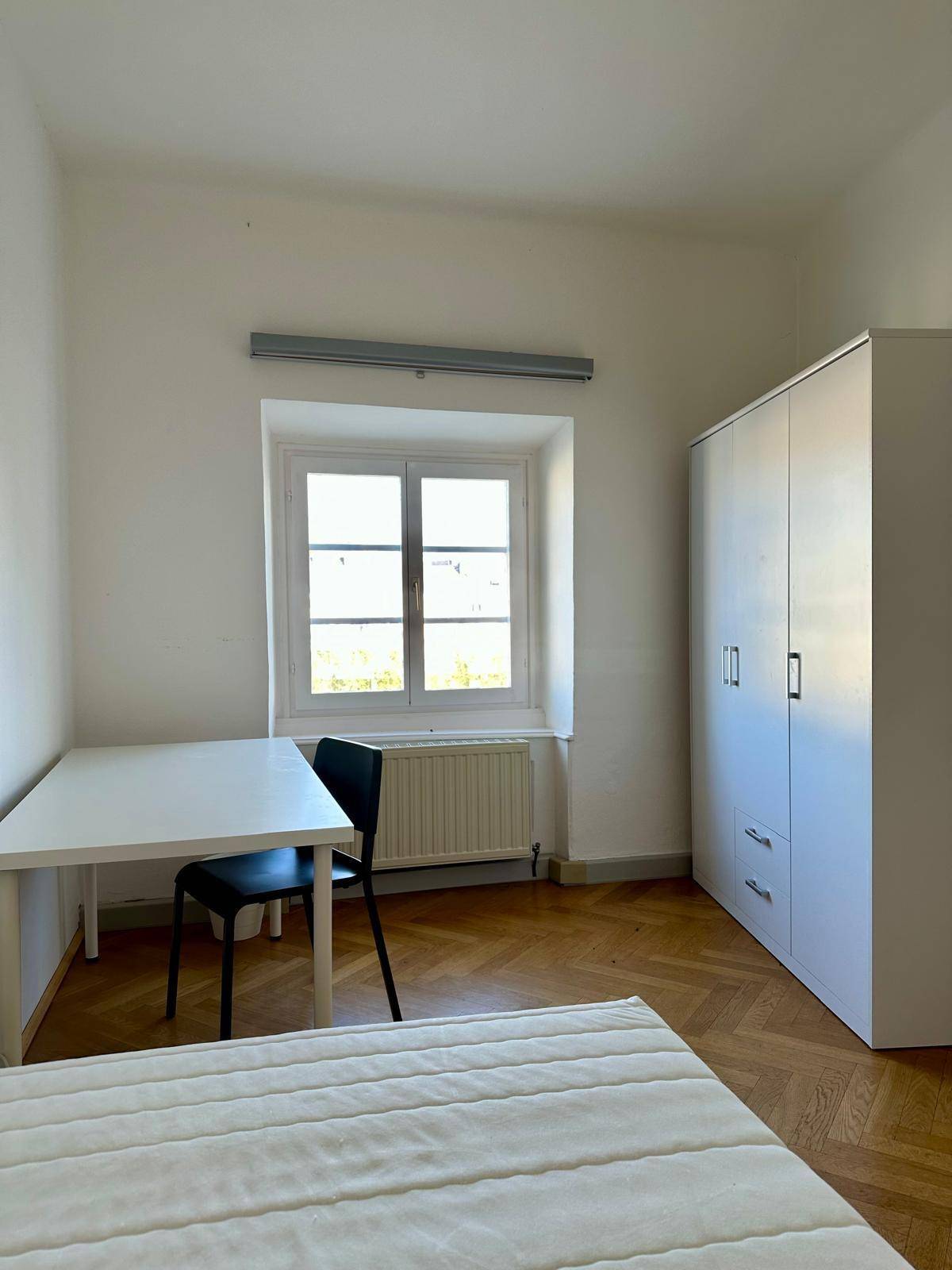 Helles Schlafzimmer mit Parkettboden, weißem Kleiderschrank und Schreibtisch am Fenster.
