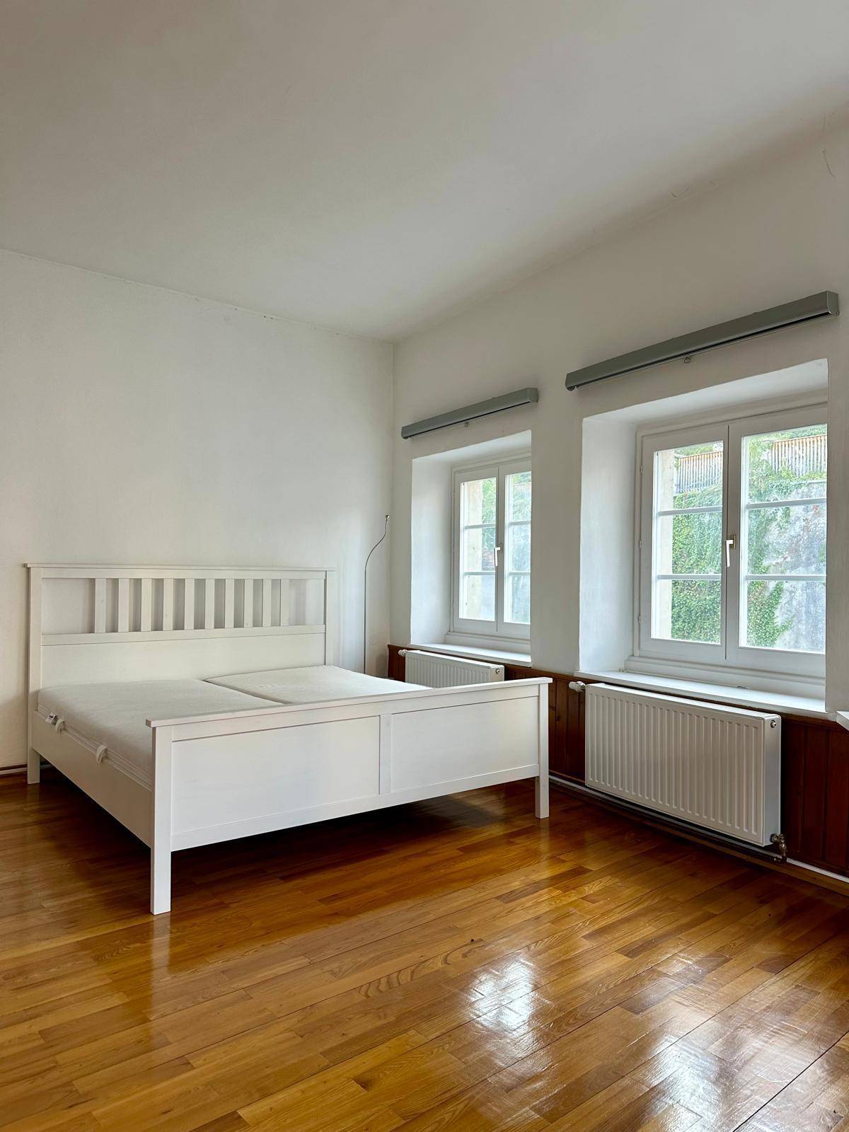 Geräumiges Schlafzimmer mit zwei Fenstern, Holzboden und einem weißen Doppelbett.