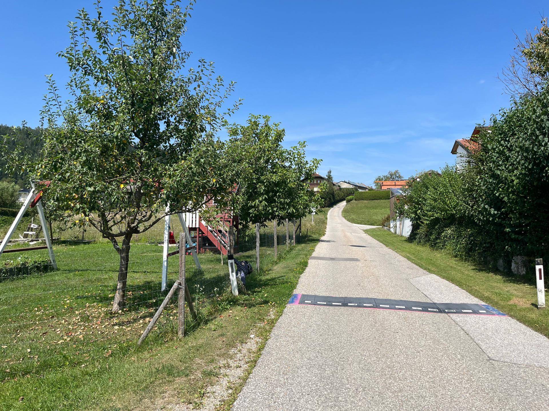 Ländliche Straße neben einem Garten mit Spielgeräten und Obstbäumen unter blauem Himmel.