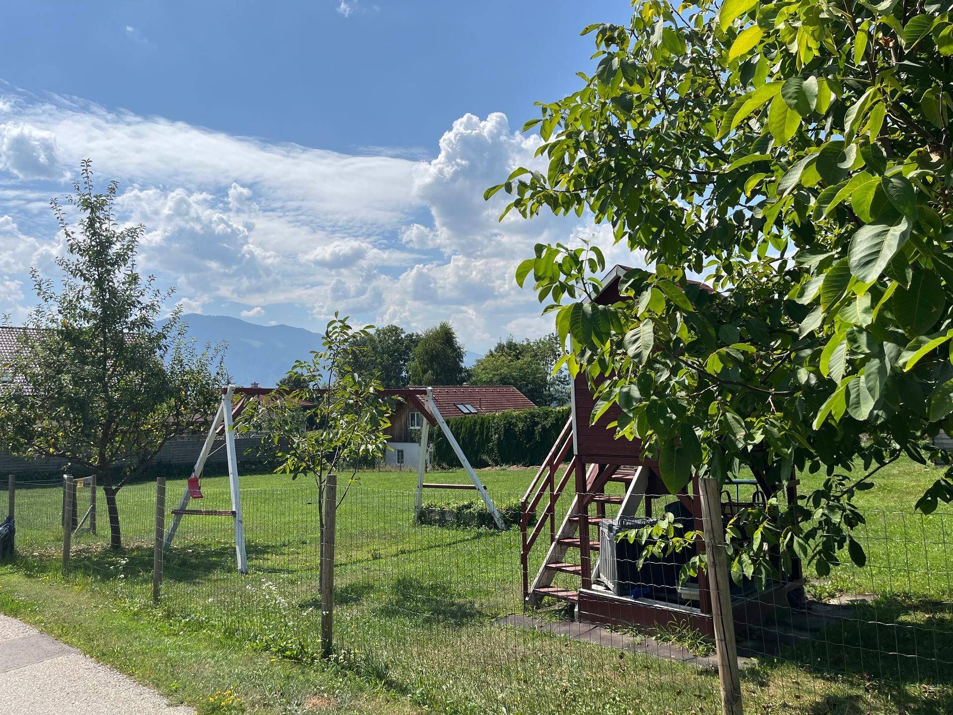 Sonniges Gartengrundstück mit Schaukel und Spielturm, umgeben von Obstbäumen und grüner Natur.