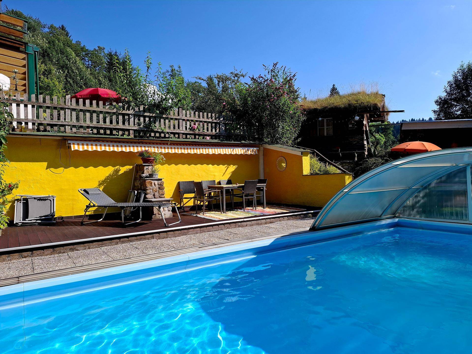 Großer Außenpool mit Schiebeabdeckung, umgeben von einer Terrasse mit Sitzgelegenheiten und Bergblick.