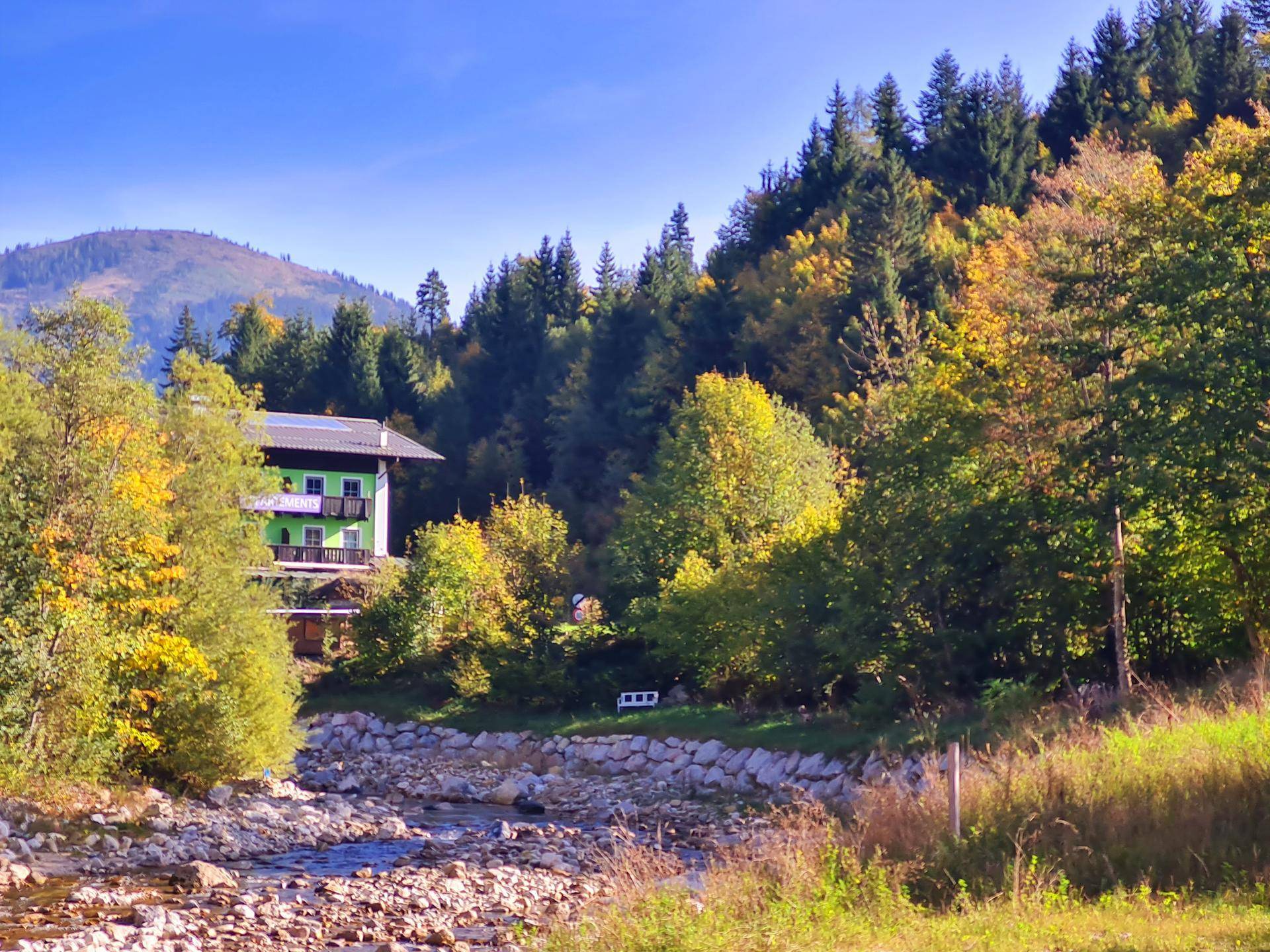 Grünes Gebäude am Flussufer, umgeben von herbstlicher Natur und bewaldeten Bergen.