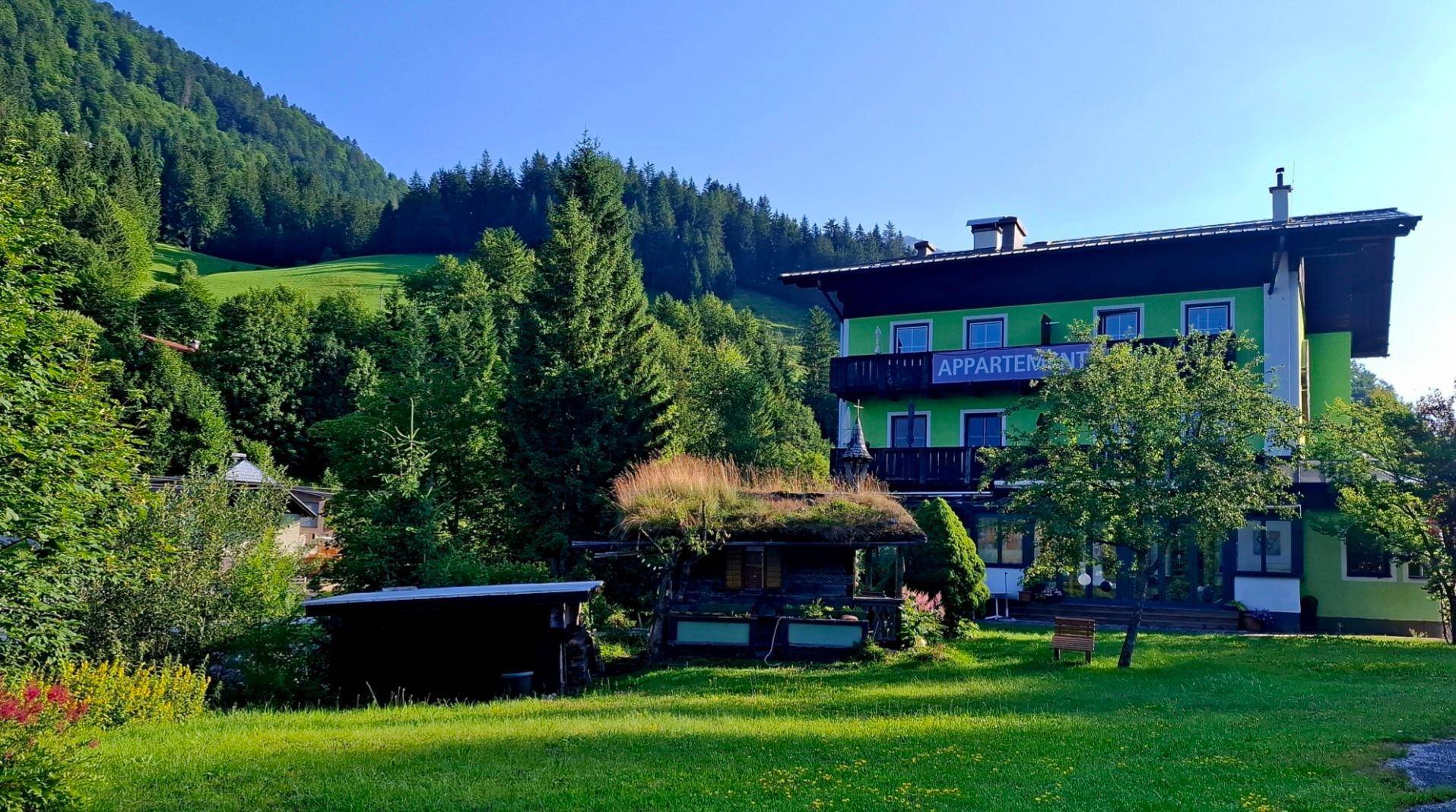 Grünes Gebäude mit Balkonen und großem Garten, eingebettet in eine malerische Berglandschaft.