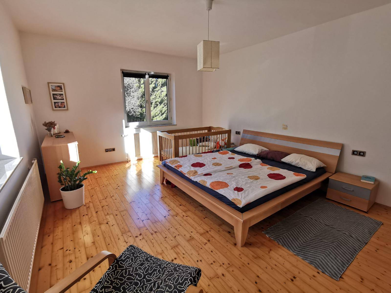 Helles Schlafzimmer mit Holzboden, Doppelbett, Babybett und Blick ins Grüne.