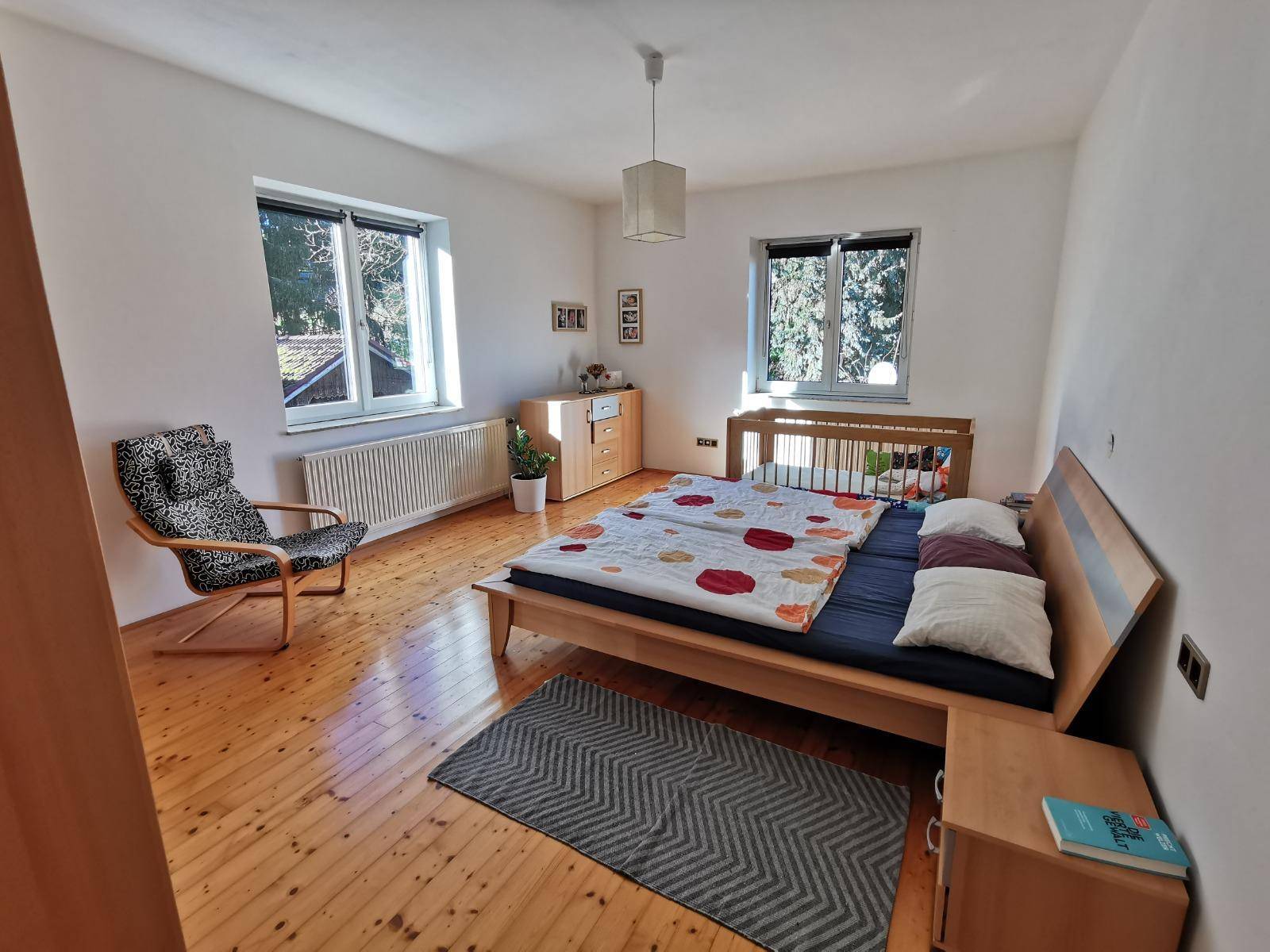 Schlafzimmer mit zwei Fenstern, Holzboden, Doppelbett und Babybett.