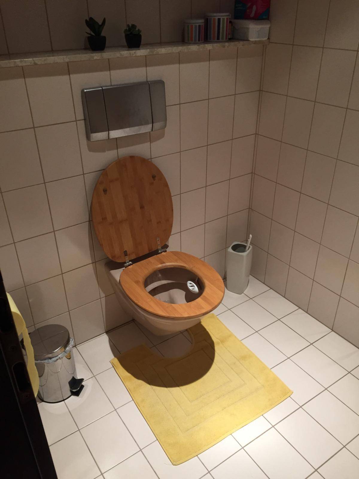 Kleines WC mit weißen Fliesen, Holz-Toilettensitz und gelbem Teppich.