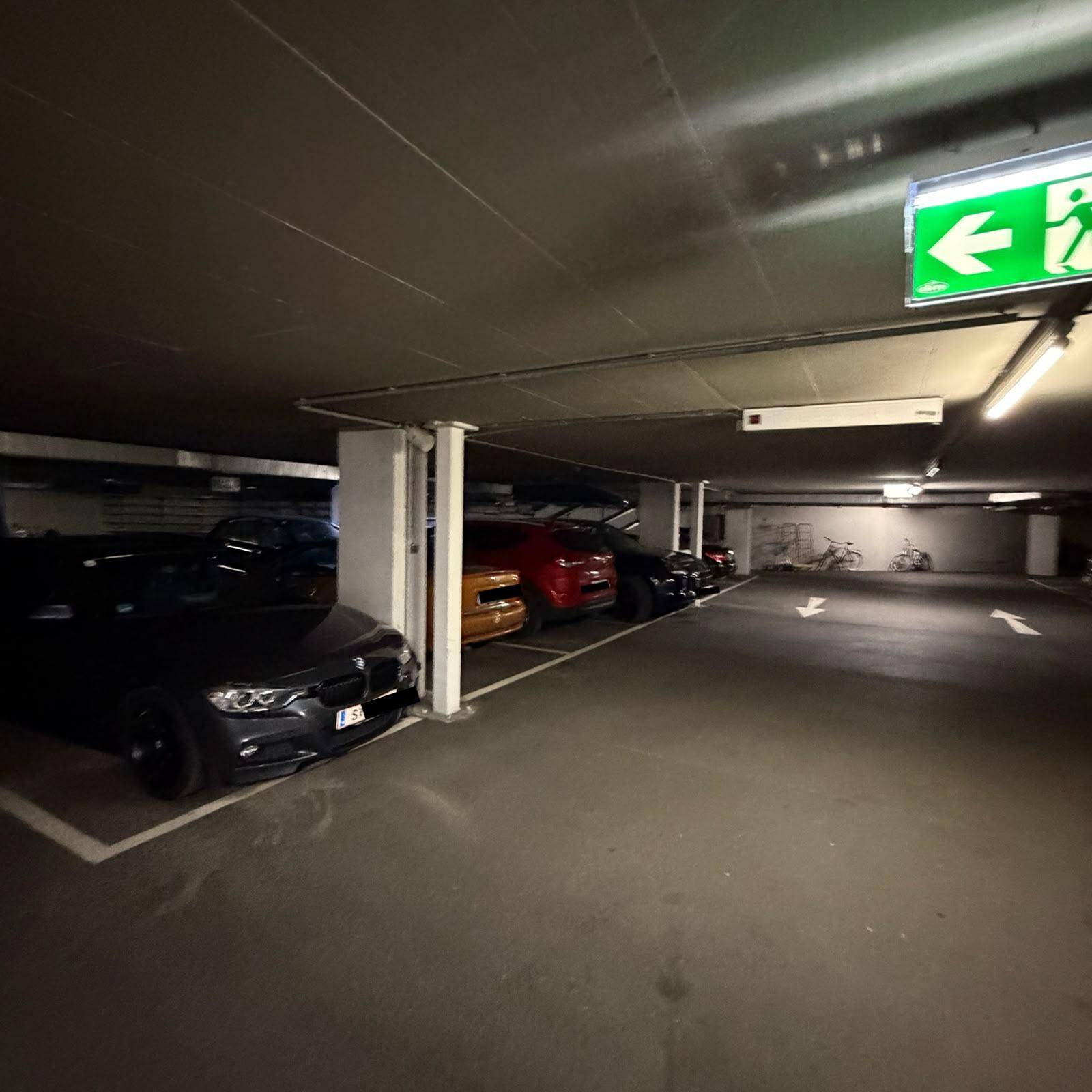 Weitläufige Tiefgarage mit geparkten Autos und einem beleuchteten Notausgangsschild an der Decke.