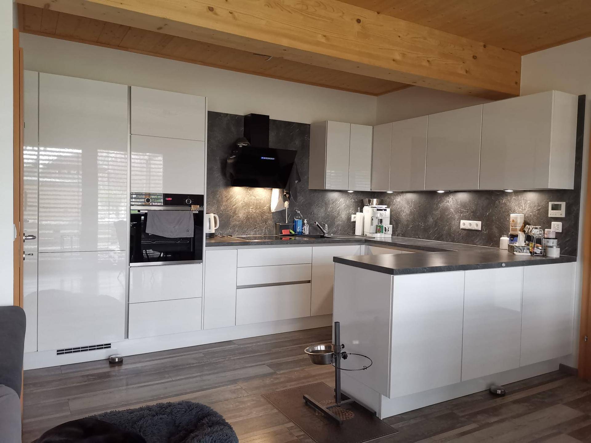 Moderne Küche mit weißen Hochglanzfronten, dunkler Arbeitsplatte und Holzbalken.
