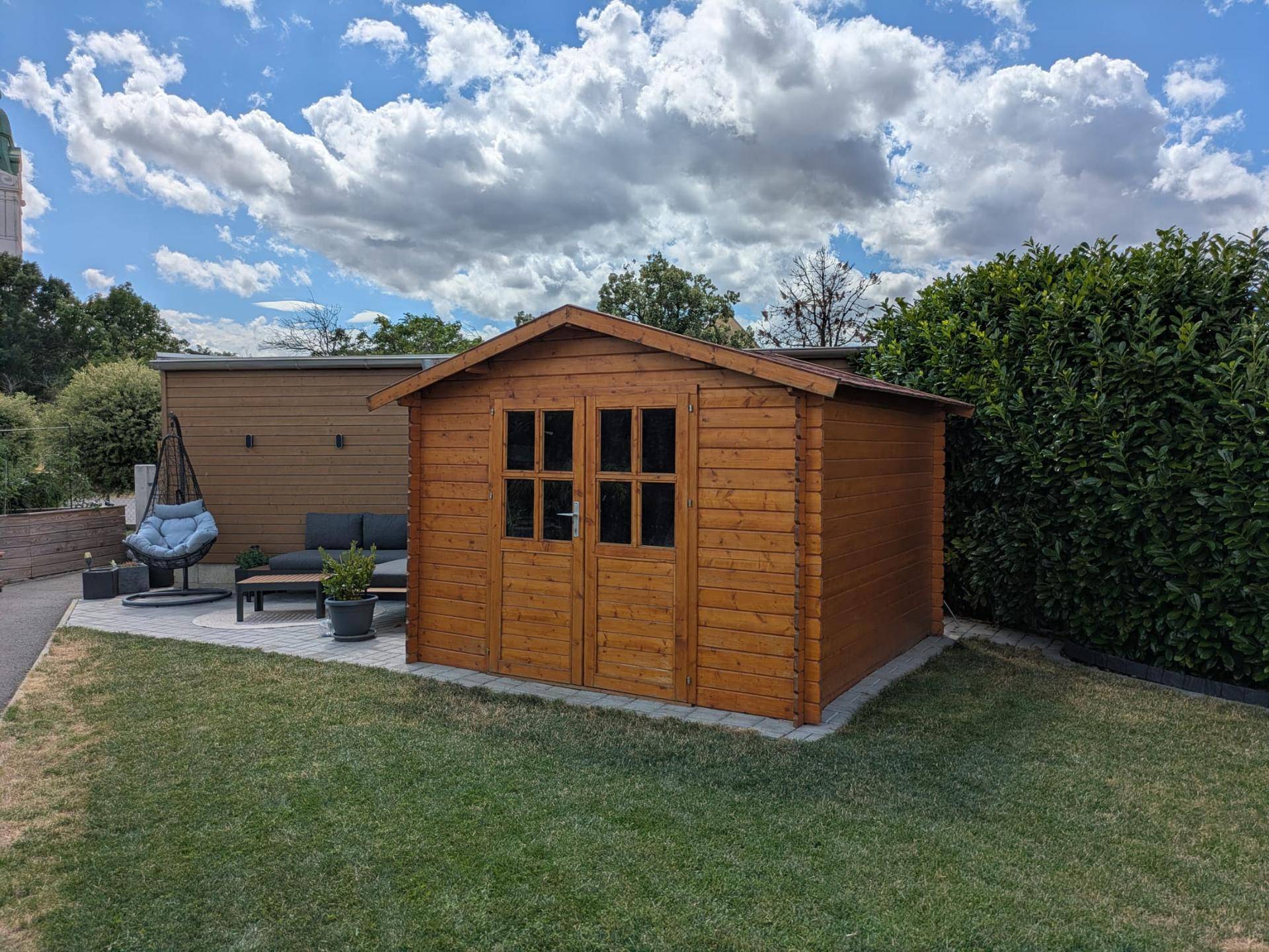 Kleiner Holzschuppen im Garten neben einer gepflasterten Terrasse mit Sitzgelegenheiten.