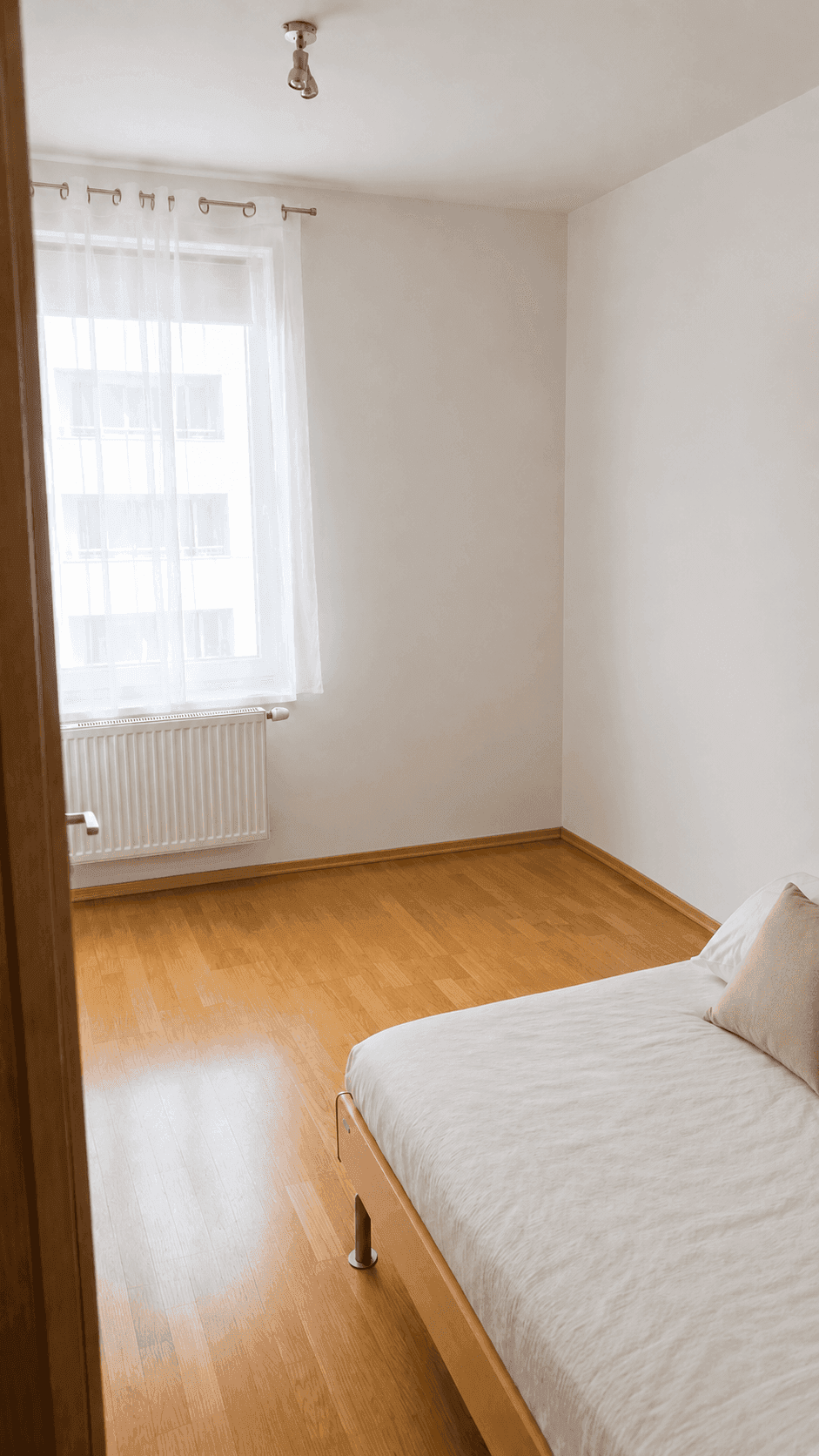 Schlichtes Schlafzimmer mit Holzboden, großem Fenster und viel natürlichem Licht.