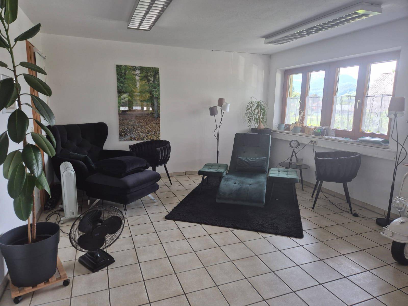 Heller Raum mit beigen Fliesenboden, bequemen Lounge-Möbeln und einem großen Fenster mit Holzjalousien.