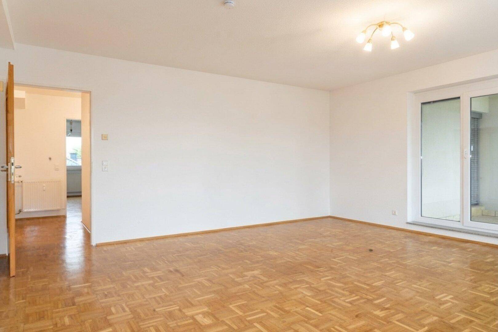 Heller Wohnbereich mit Parkettboden und Fensterfront, die viel Tageslicht hereinlässt.
