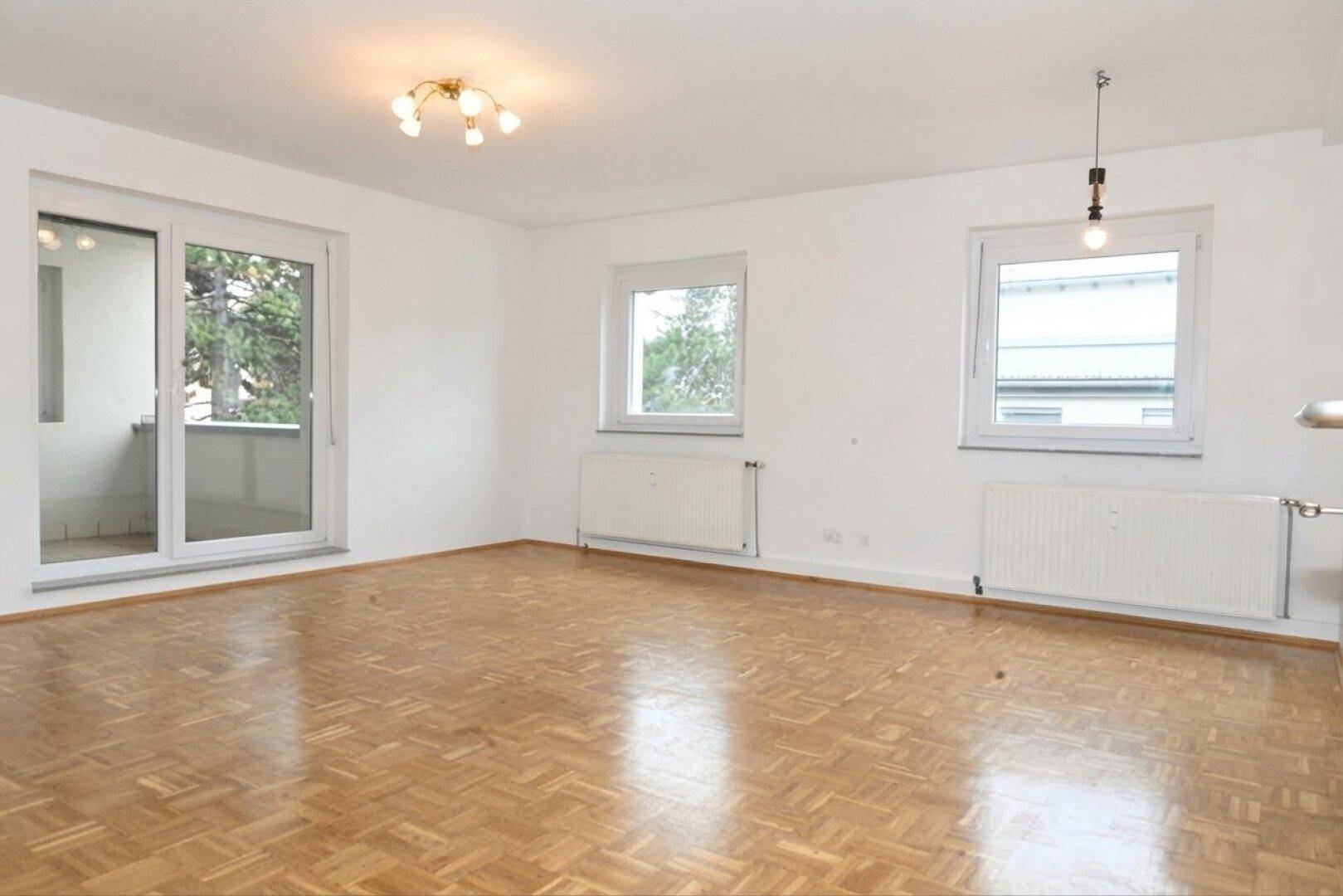 Helles Wohnzimmer mit Parkettboden, Heizkörpern und Zugang zu einem Balkon.