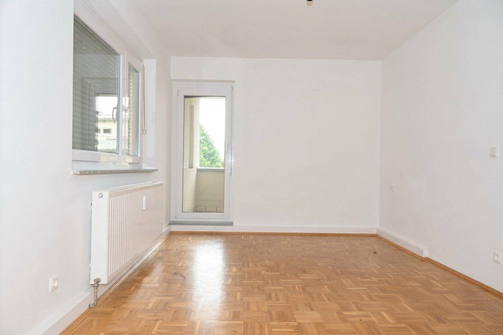 Helles Schlafzimmer mit Parkettboden, Fenster und Zugang zu einem kleinen Balkon.