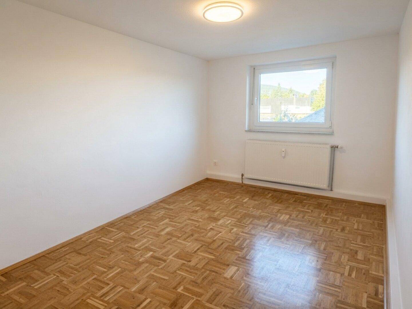 Helles Schlafzimmer mit Parkettboden, großem Fenster und Heizkörper.