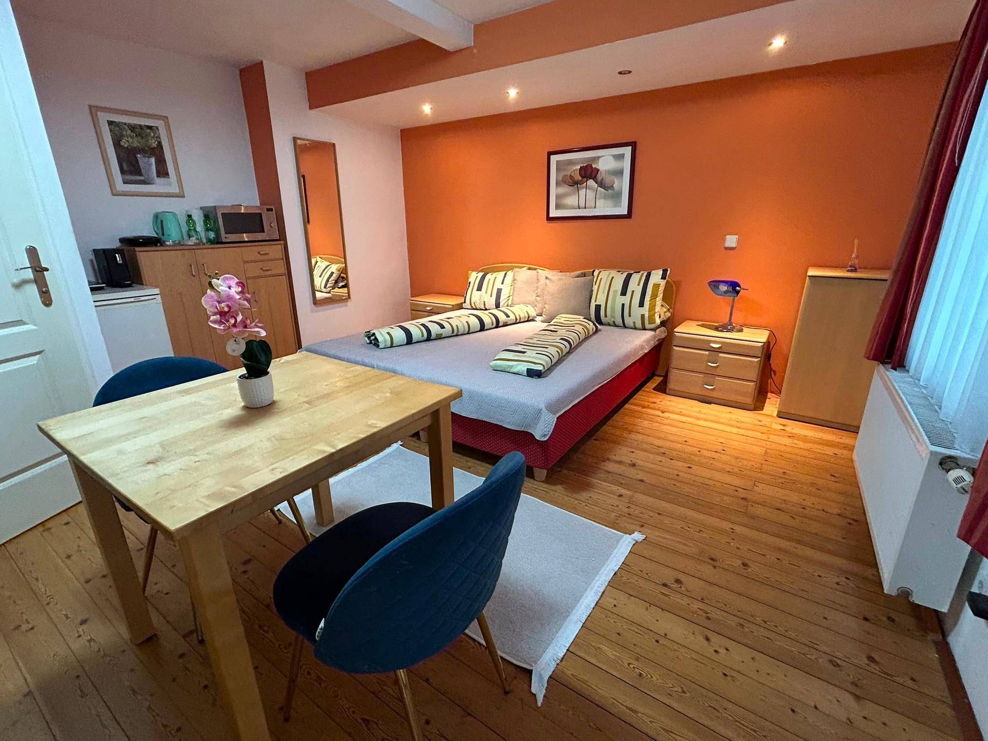 Geräumiges Studio-Apartment mit Doppelbett, Essbereich und Holzboden in warmen Orangetönen.