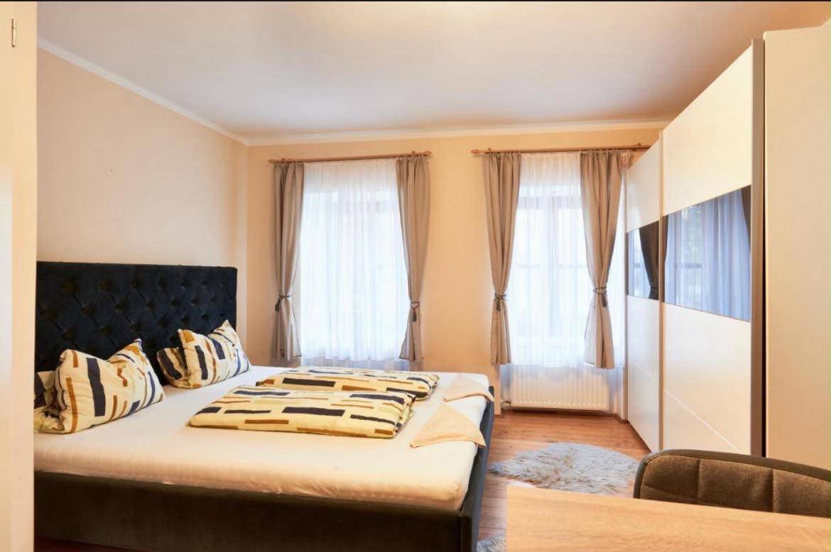 Helles Schlafzimmer mit Doppelbett, großem Kleiderschrank und zwei Fenstern mit Vorhängen.
