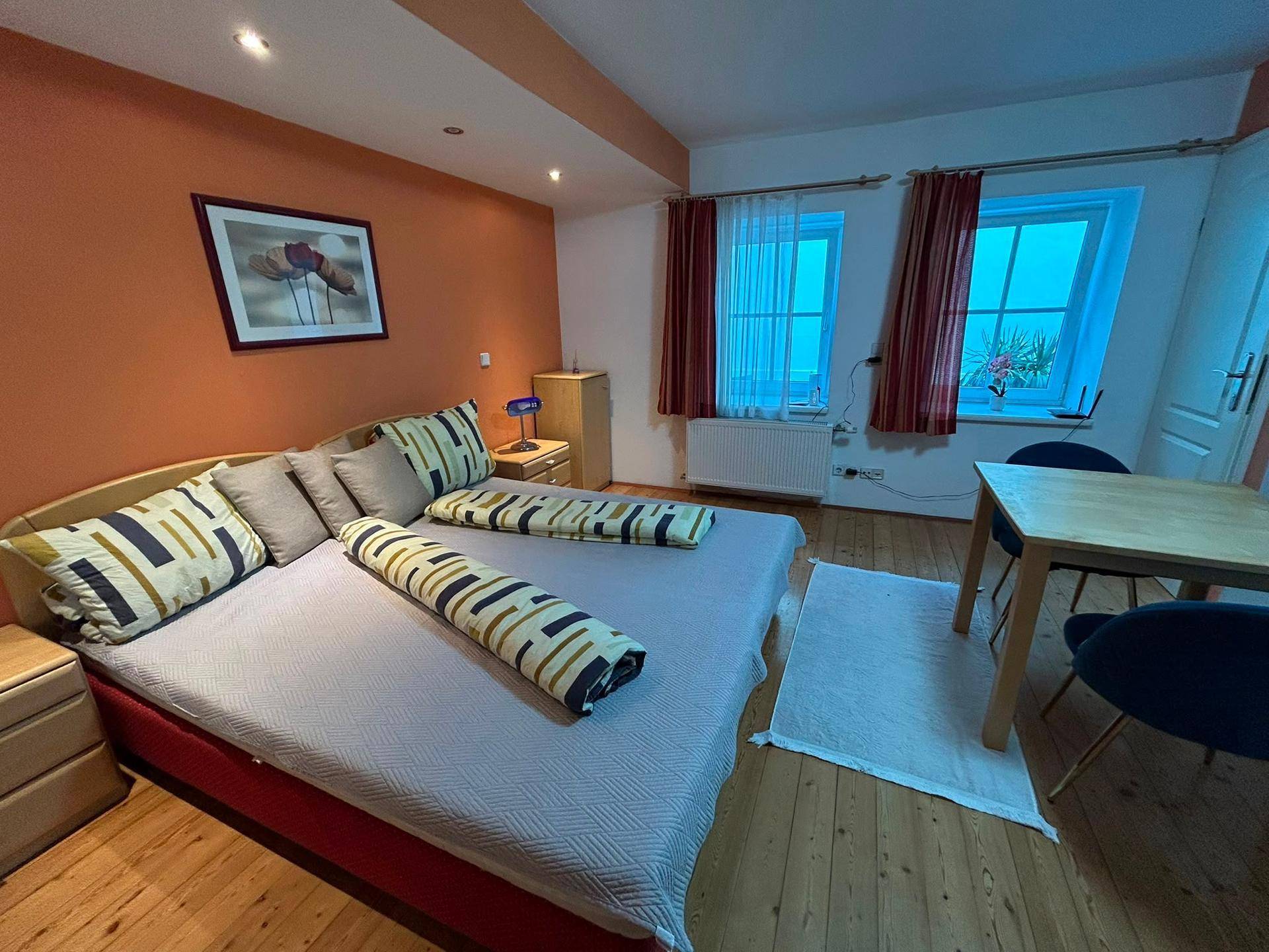 Geräumiges Studio-Apartment mit Doppelbett, Essbereich und Holzboden in warmen Orangetönen.