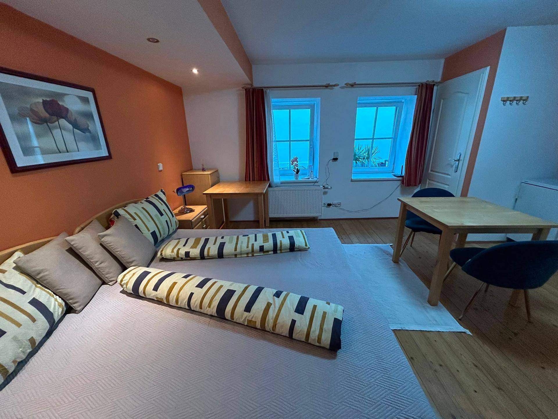 Geräumiges Studio-Apartment mit Doppelbett, Essbereich und Holzboden in warmen Orangetönen.
