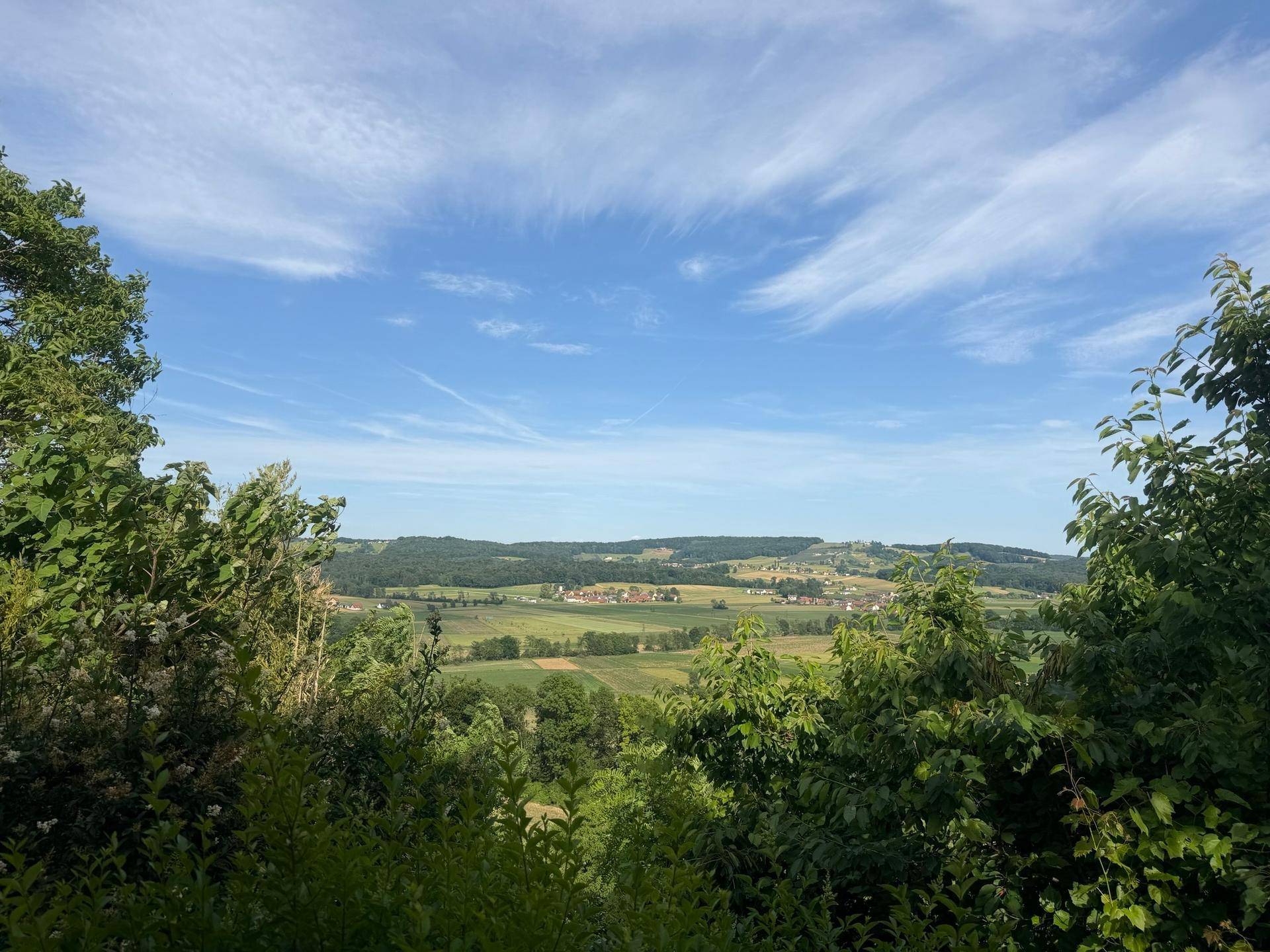 Weitläufiger Blick über eine grüne Hügellandschaft mit Feldern und kleinen Dörfern unter blauem Himmel.