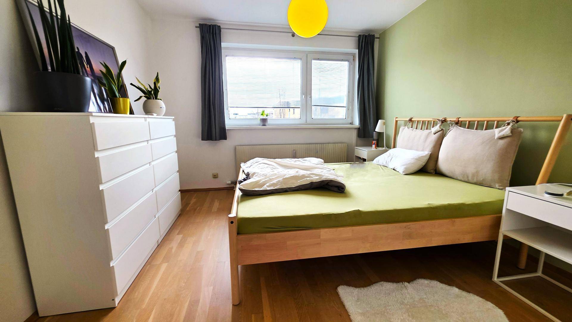 Gemütliches Schlafzimmer mit Holzbett, grüner Wand und viel Tageslicht durch das Fenster.