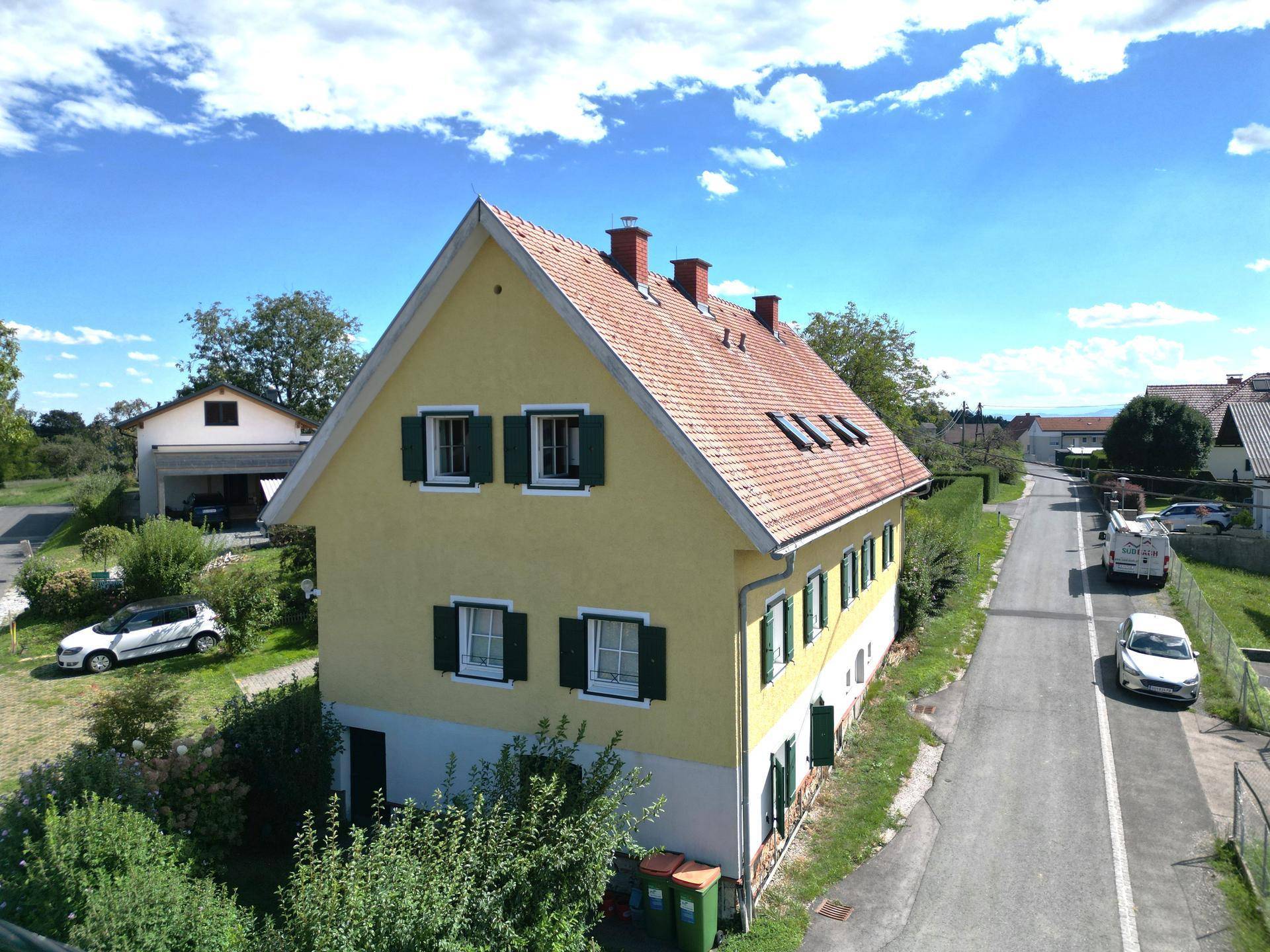 Seitenansicht eines gelben Hauses mit rotem Ziegeldach und grünen Fensterläden.