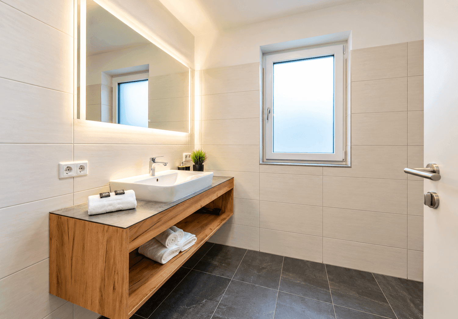 Modernes Badezimmer mit schwebendem Waschtisch, beleuchtetem Spiegel und Fenster.