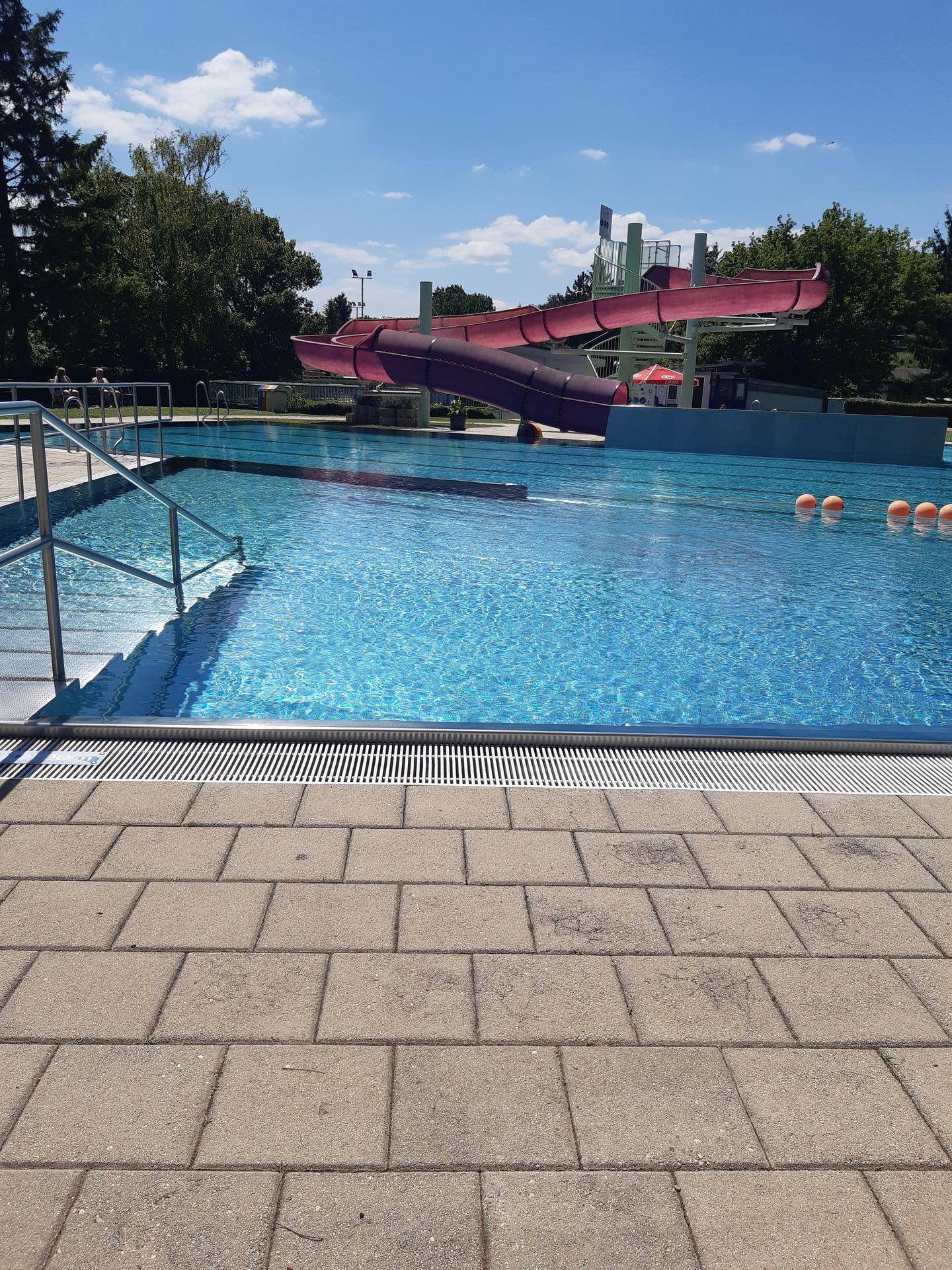 Großes Schwimmbecken mit Wasserrutschen, umgeben von Bäumen unter blauem Himmel.