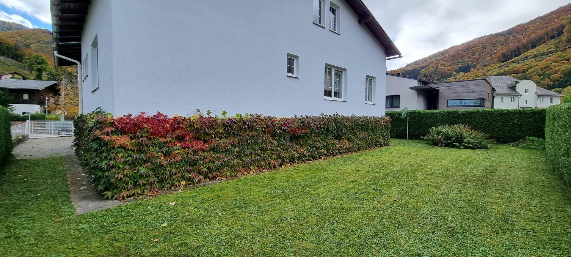 Seitenansicht des Hauses mit gepflegtem Rasen, bunter Hecke und Bergblick.