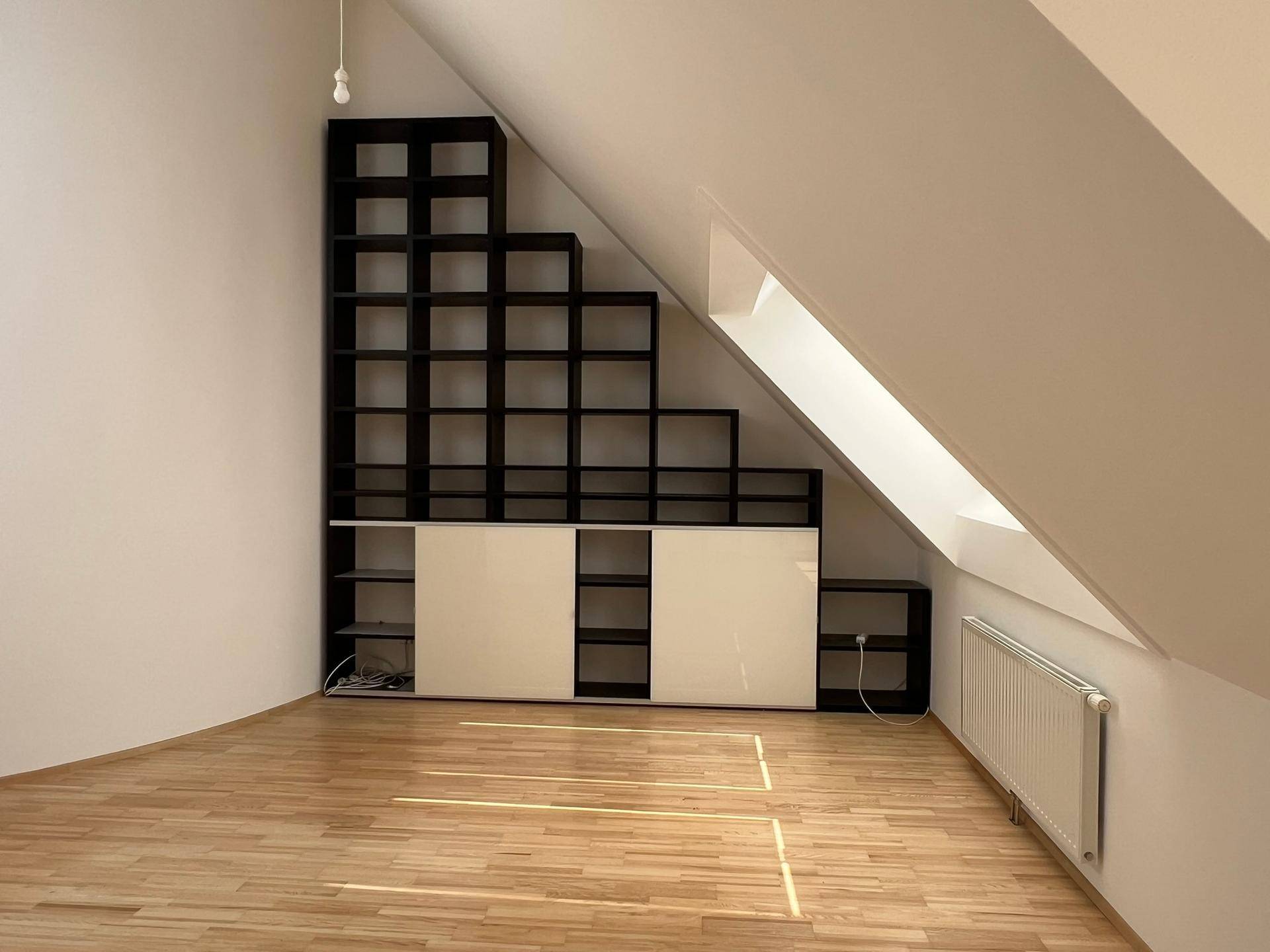 Dachzimmer mit schrägen Wänden, Holzboden und einem integrierten, stufenförmigen Regalsystem.