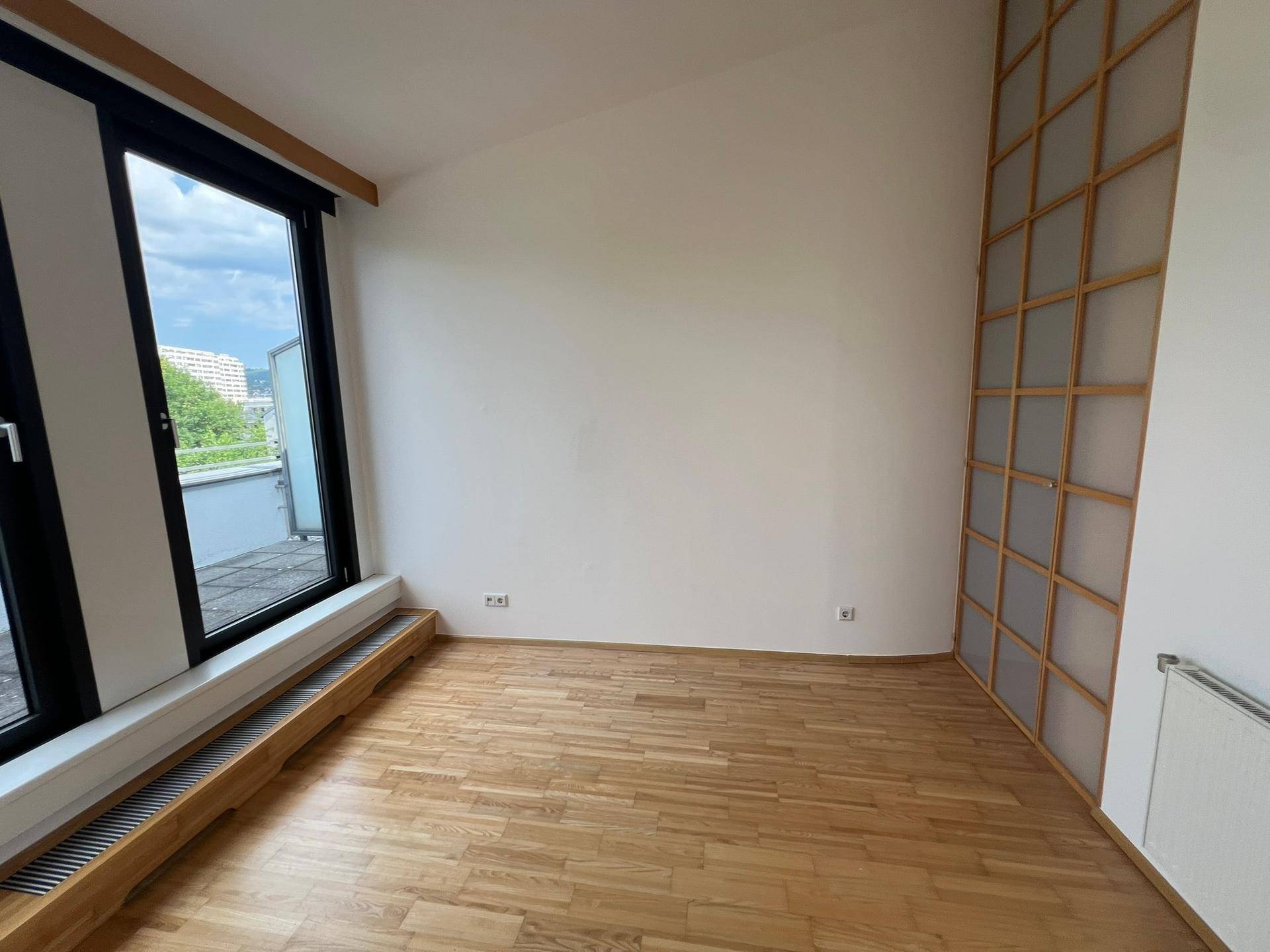 Helles Zimmer mit Holzboden, großem Fenster und einer dekorativen Holztrennwand.
