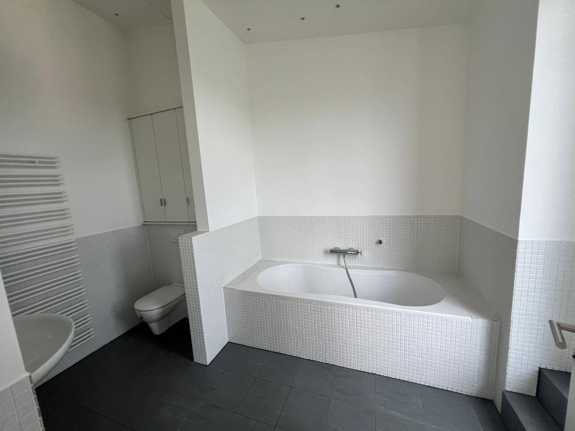 Badezimmer mit Badewanne, WC und Handtuchheizkörper, gefliest in Weiß und Grau.