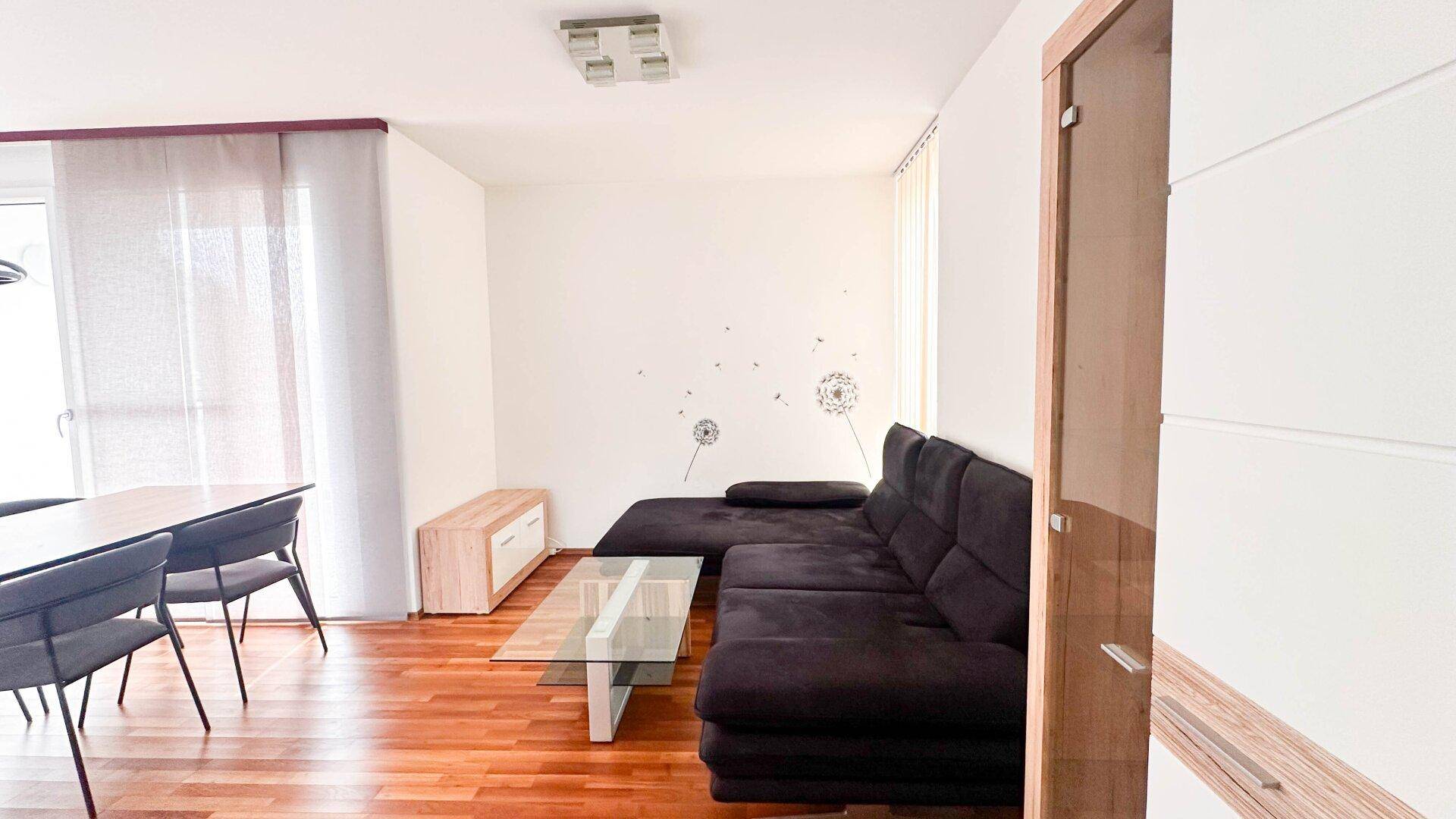 Gemütlicher Wohnbereich mit dunklem Ecksofa und Couchtisch, verbunden mit dem Essbereich.