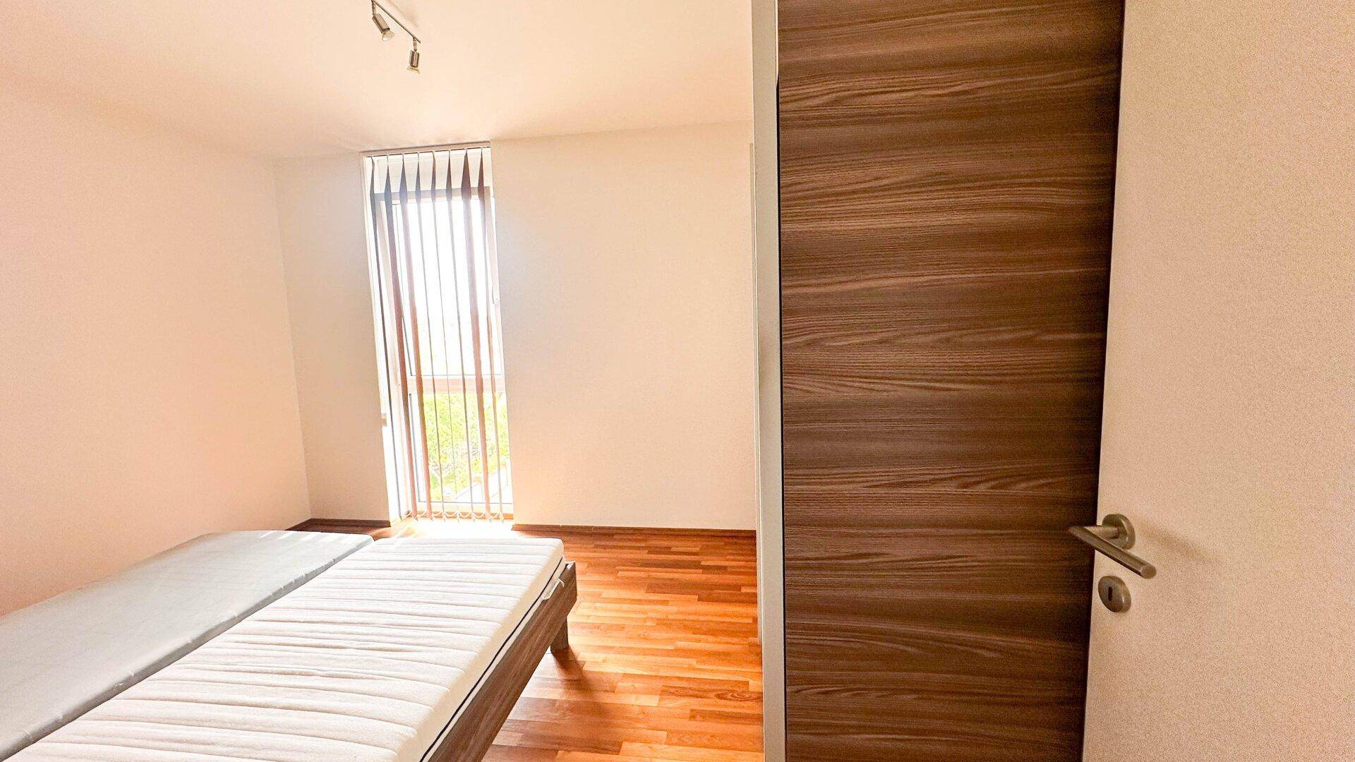 Helles Schlafzimmer mit Holzboden und großem Fenster, das viel Tageslicht hereinlässt.