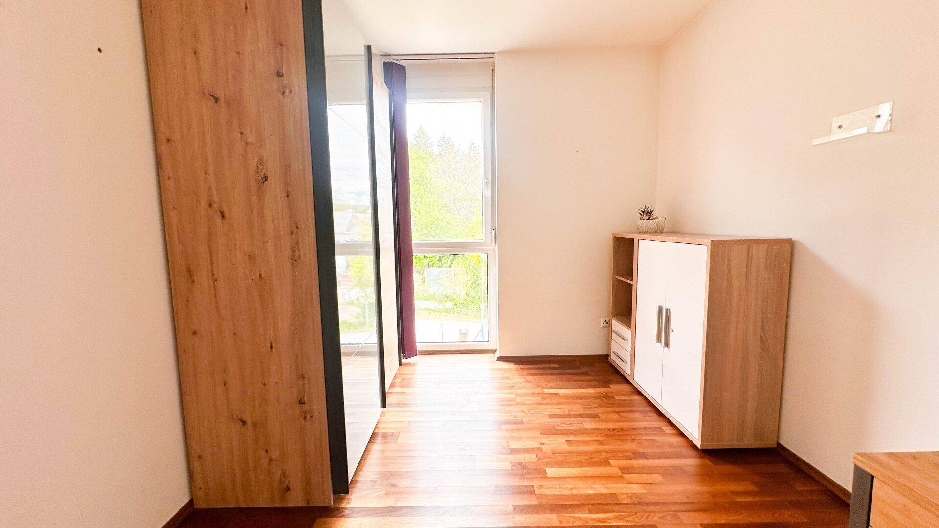 Helles Zimmer mit Holzboden und großem Fenster, ausgestattet mit einem modernen Kleiderschrank.