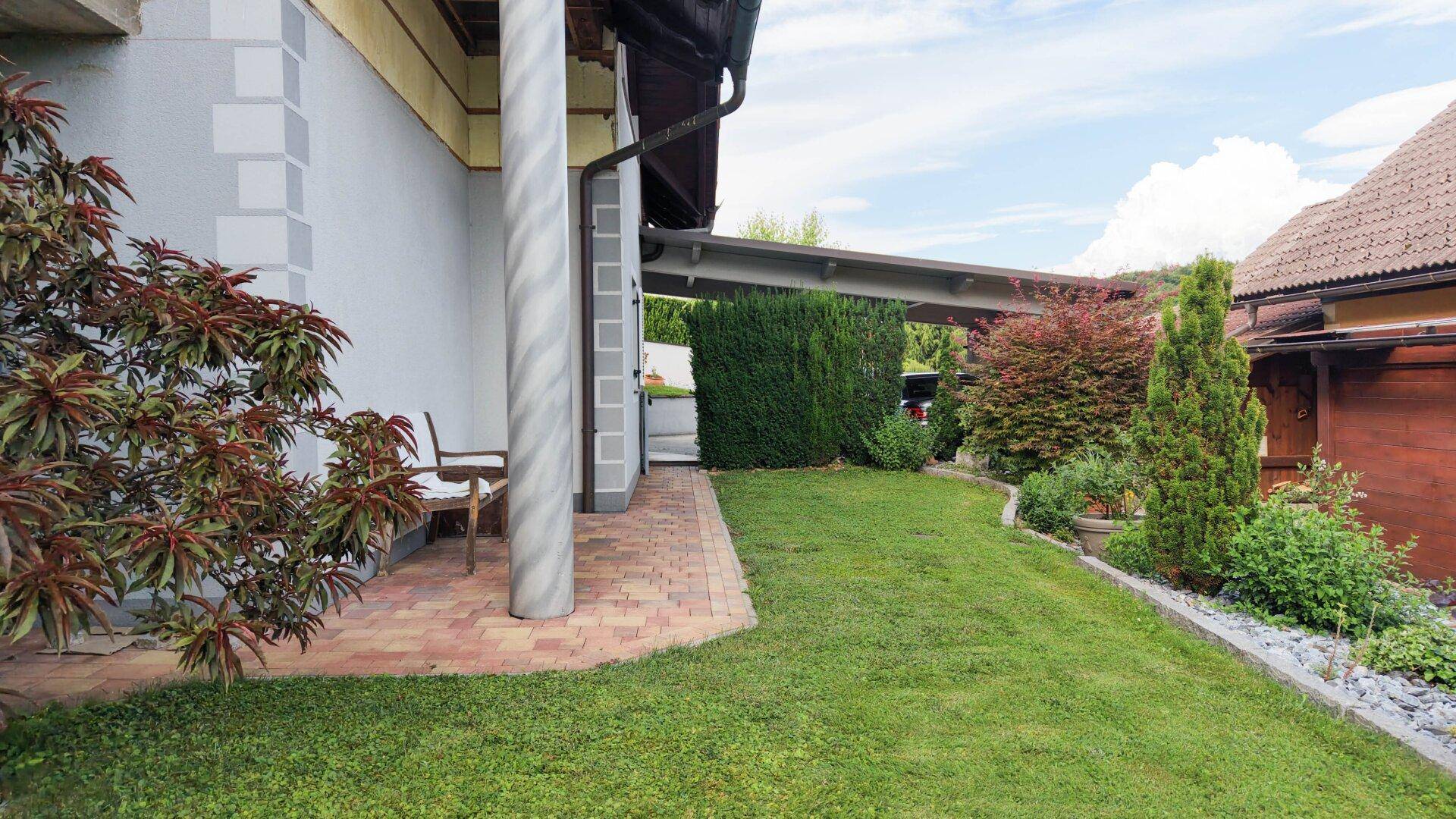Gepflegte Rasenfläche neben dem Haus mit gepflastertem Weg und Carport im Hintergrund.