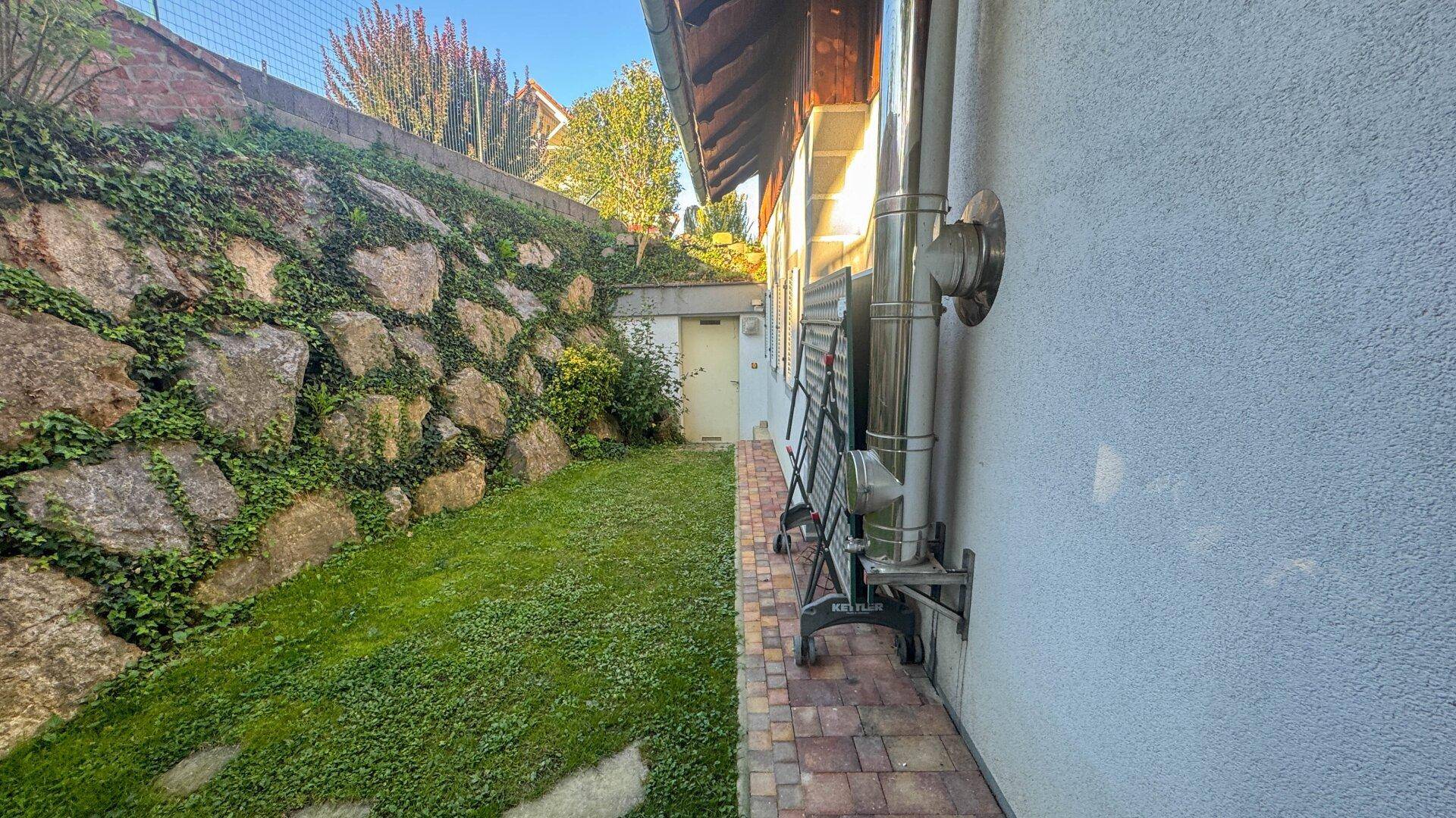 Schmaler Gartenbereich neben dem Haus mit einer begrünten Steinmauer und Rasen.