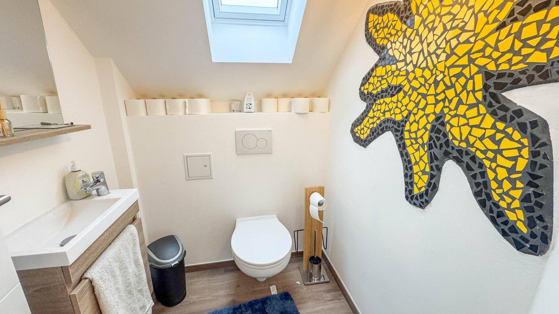 Modernes WC mit Dachfenster, Wand-WC und einem auffälligen gelben Mosaikbild.