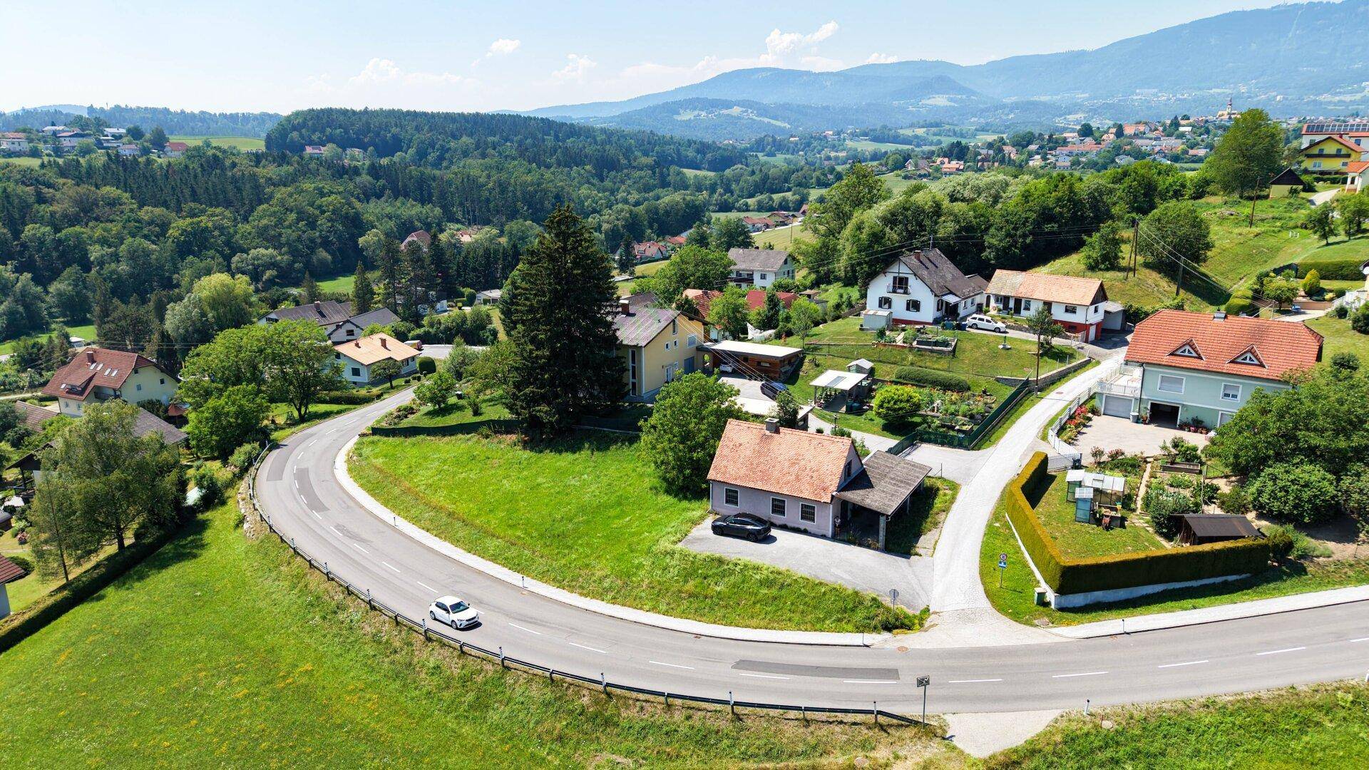 Weitläufige Luftaufnahme einer Wohnsiedlung in ländlicher Umgebung mit kurviger Straße und grüner Landschaft.