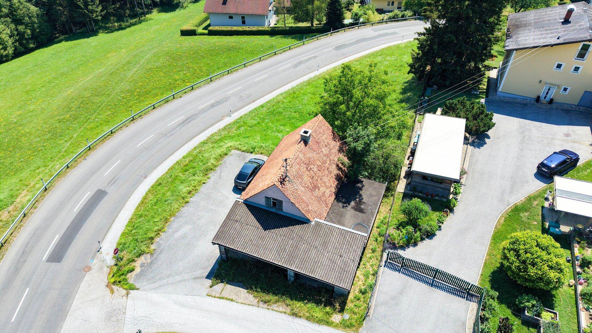 Draufsicht auf ein Wohnhaus mit Ziegeldach und einem langen Carport, umgeben von Grünflächen und einer Straße.