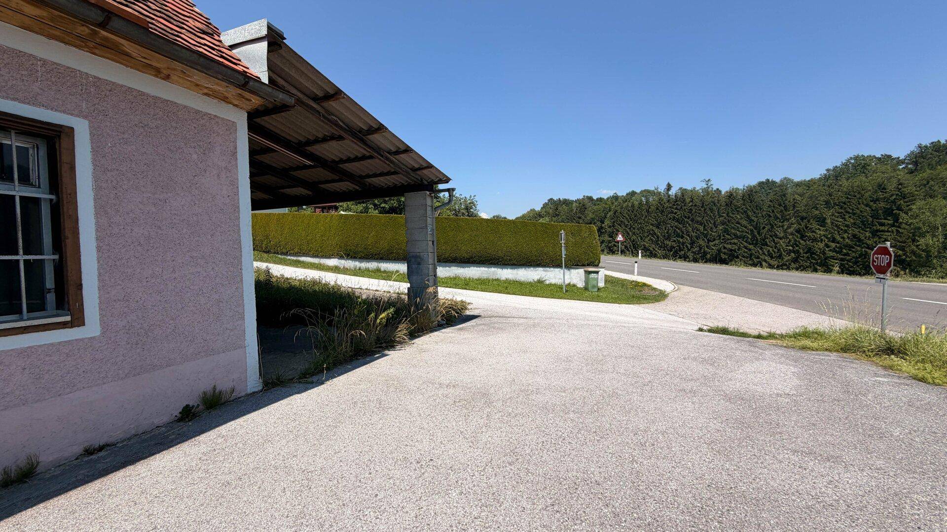 Seitenansicht des Hauses mit Carport und Zufahrt, im Hintergrund eine Straße und grüne Hecken.