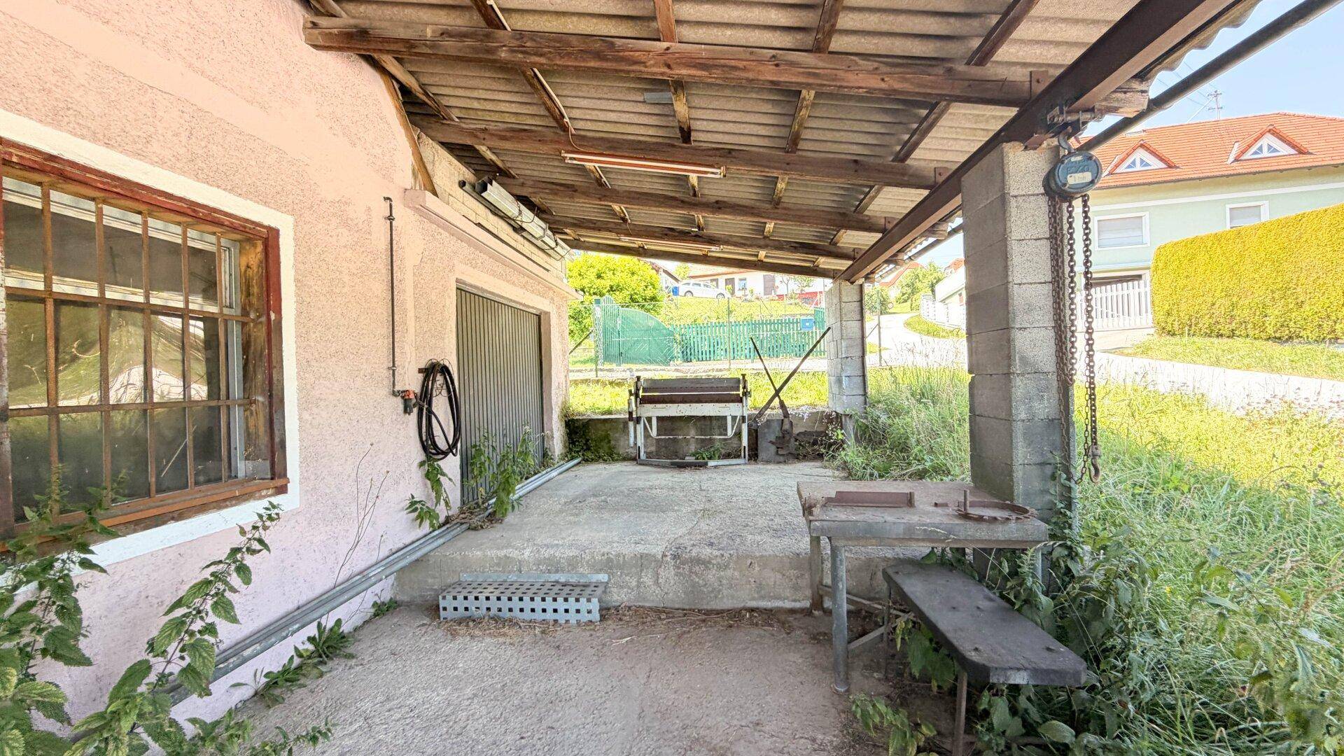 Überdachter Außenbereich mit Werkbank und Geräten, angrenzend an die Hauswand mit Fenster.
